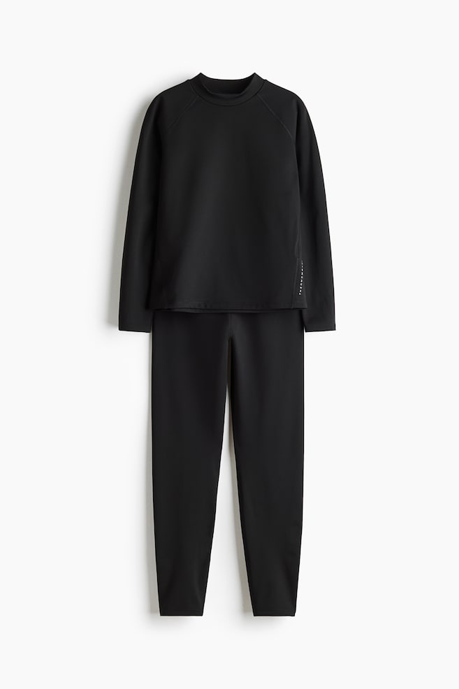A black two-piece activewear set, front-facing, displaying a long-sleeve mock-neck top and matching slim-fit trousers. The top features raglan sleeves with a small vertical white text detail on the lower left side, while the trousers show a tapered-leg silhouette. - 1