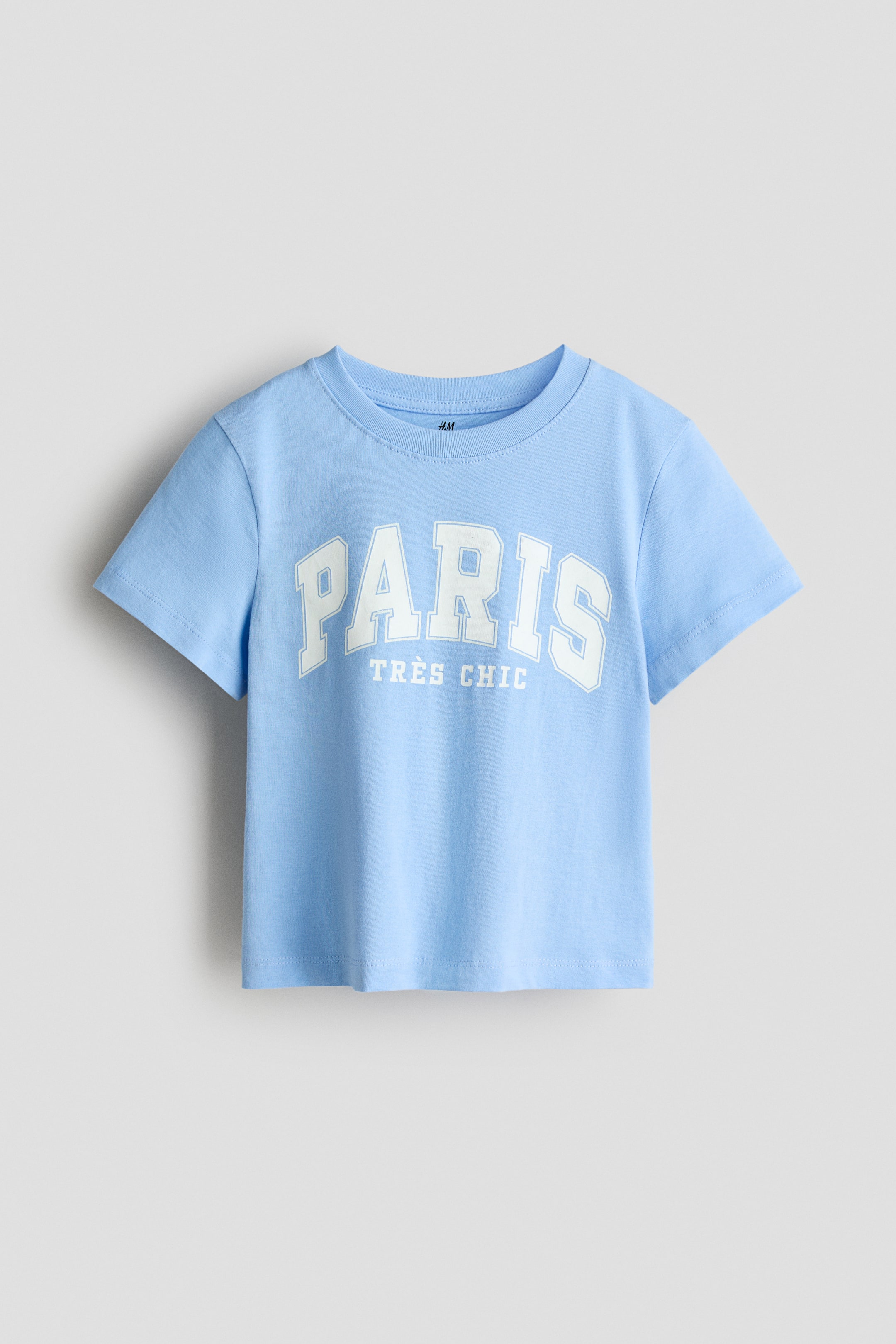 View larger image: A light blue jersey t-shirt is showcasing its front view, featuring a white printed graphic across the chest that reads 'PARIS' in large, arched letters above 'TRÈS CHIC' in smaller letters. It is designed with a round neckline showcasing a ribbed trim.