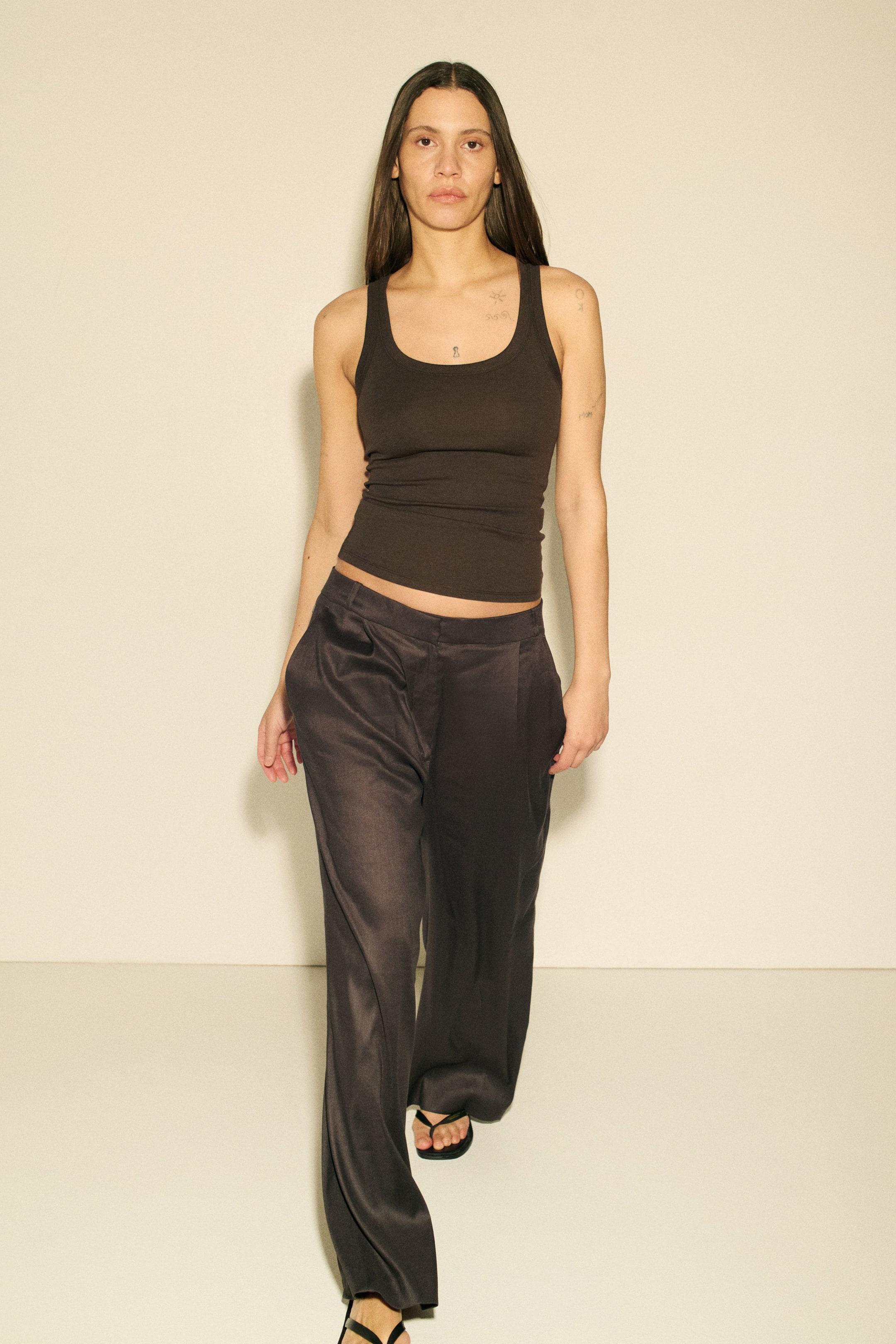 View larger image: A dark brown tank top with a scoop neck is paired with dark grey, wide-leg trousers, shown from a slightly angled front perspective. The trousers display a flowing, subtly shiny fabric with side pockets, extending over black thong sandals.
