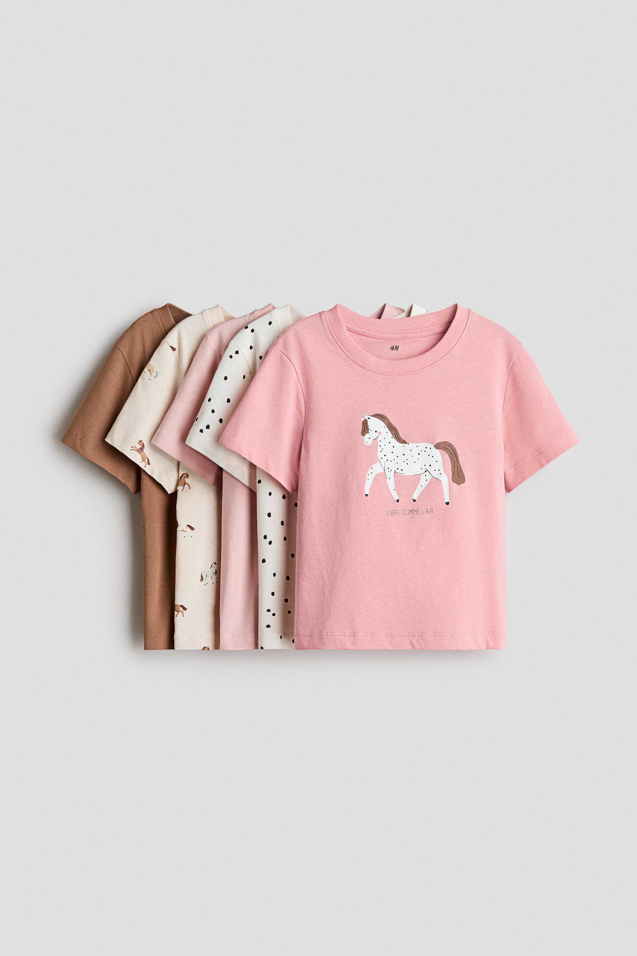 View larger image: Five short-sleeved children's T-shirts are arranged in a layered row, overlapping and facing forward. The front-most light pink T-shirt features a crew neck and a graphic of a white spotted horse with the text "LIBRE COMME L'AIR" printed underneath, while the partially visible shirts behind it feature solid brown, cream with small horse prints, solid light pink, and cream with black spots.