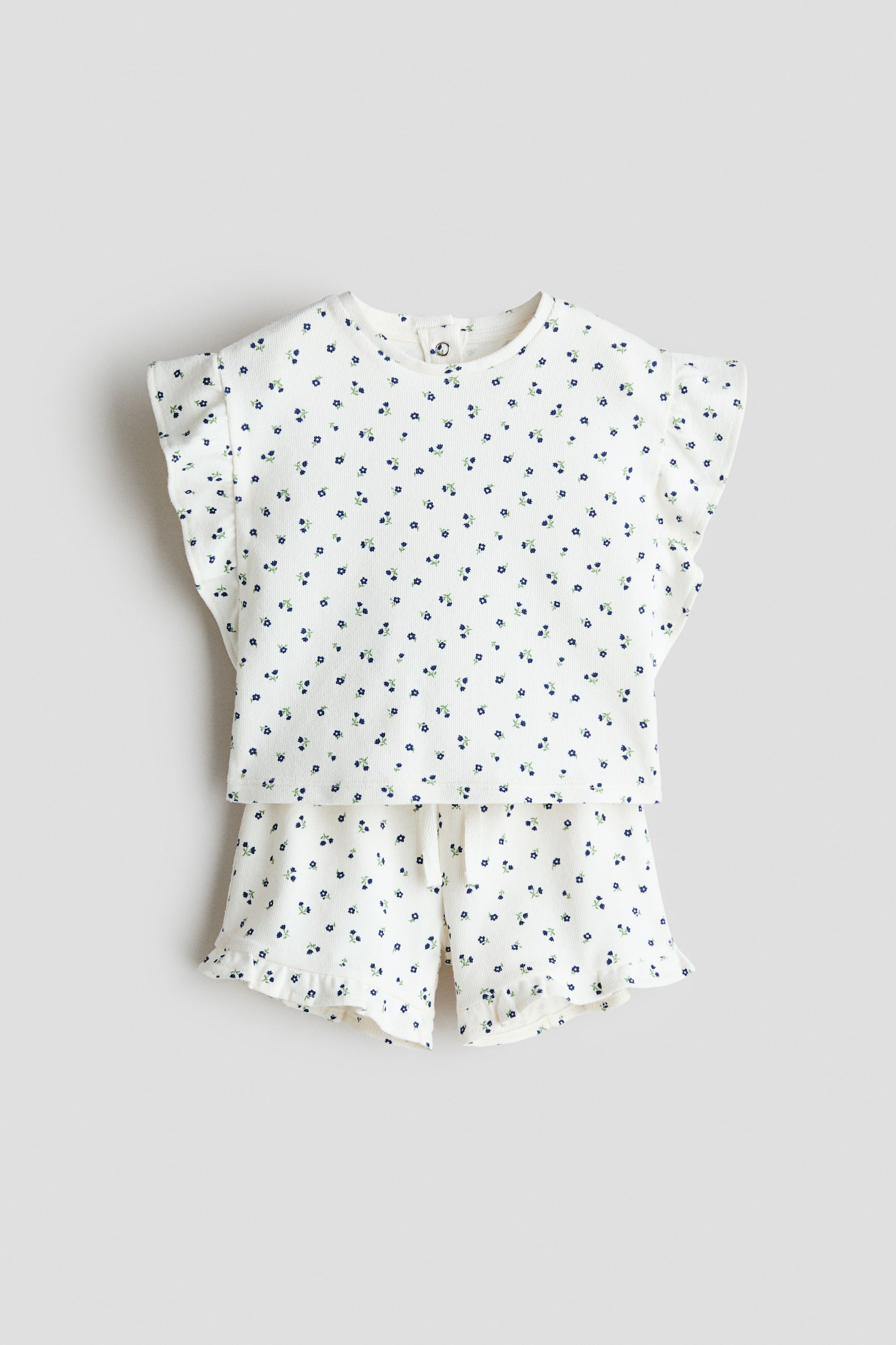 Grotere afbeelding bekijken: Een vooraanzicht van een tweedelig setje toont een off-white top met wafelstructuur, ronde hals en gerimpelde kapmouwtjes, en bijpassende shorts met een trekkoord in de taille en gerimpelde beenopeningen, allemaal versierd met een kleine blauwe bloemenprint.