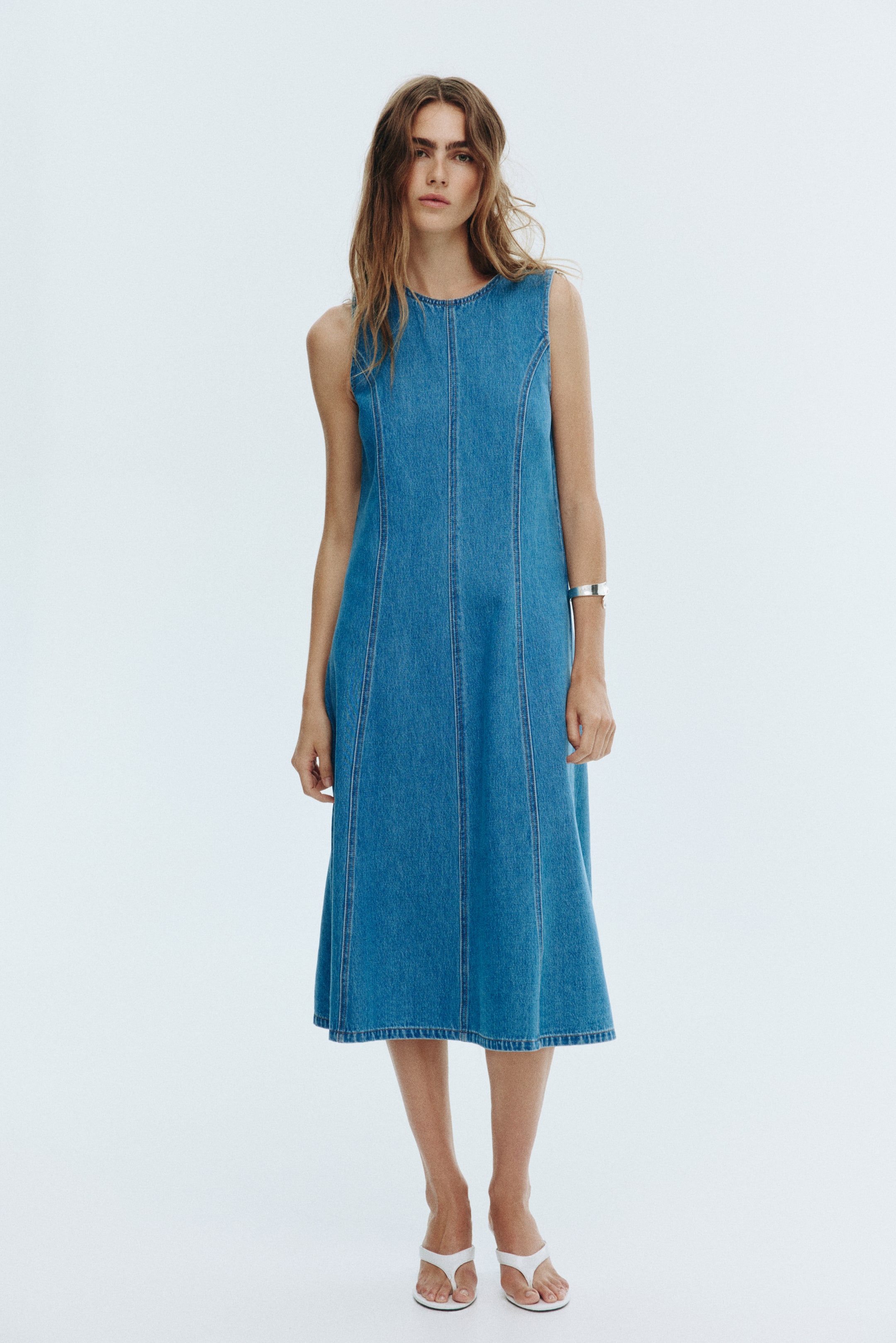 View larger image: A medium blue denim dress is being showcased, featuring a round neckline and a sleeveless cut. The dress is designed with prominent vertical seam detailing that is forming a fitted bodice and an A-line silhouette, extending to a midi length.