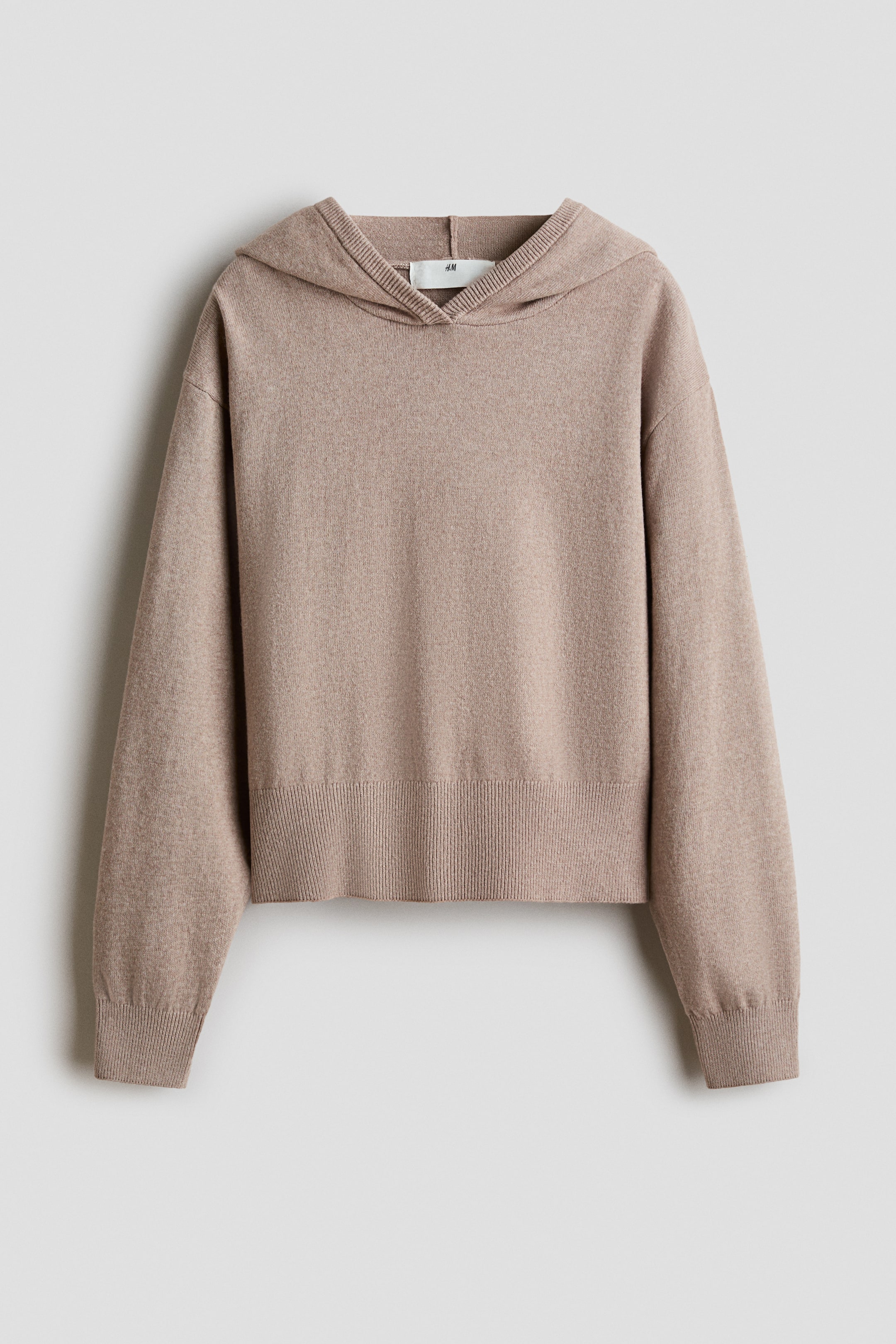View larger image: A beige fine-knit hoodie is shown from the front, featuring a relaxed fit with dropped shoulders, long sleeves, and ribbed cuffs. The garment is designed with a wide ribbed hem and a hood with a ribbed-edge neckline.