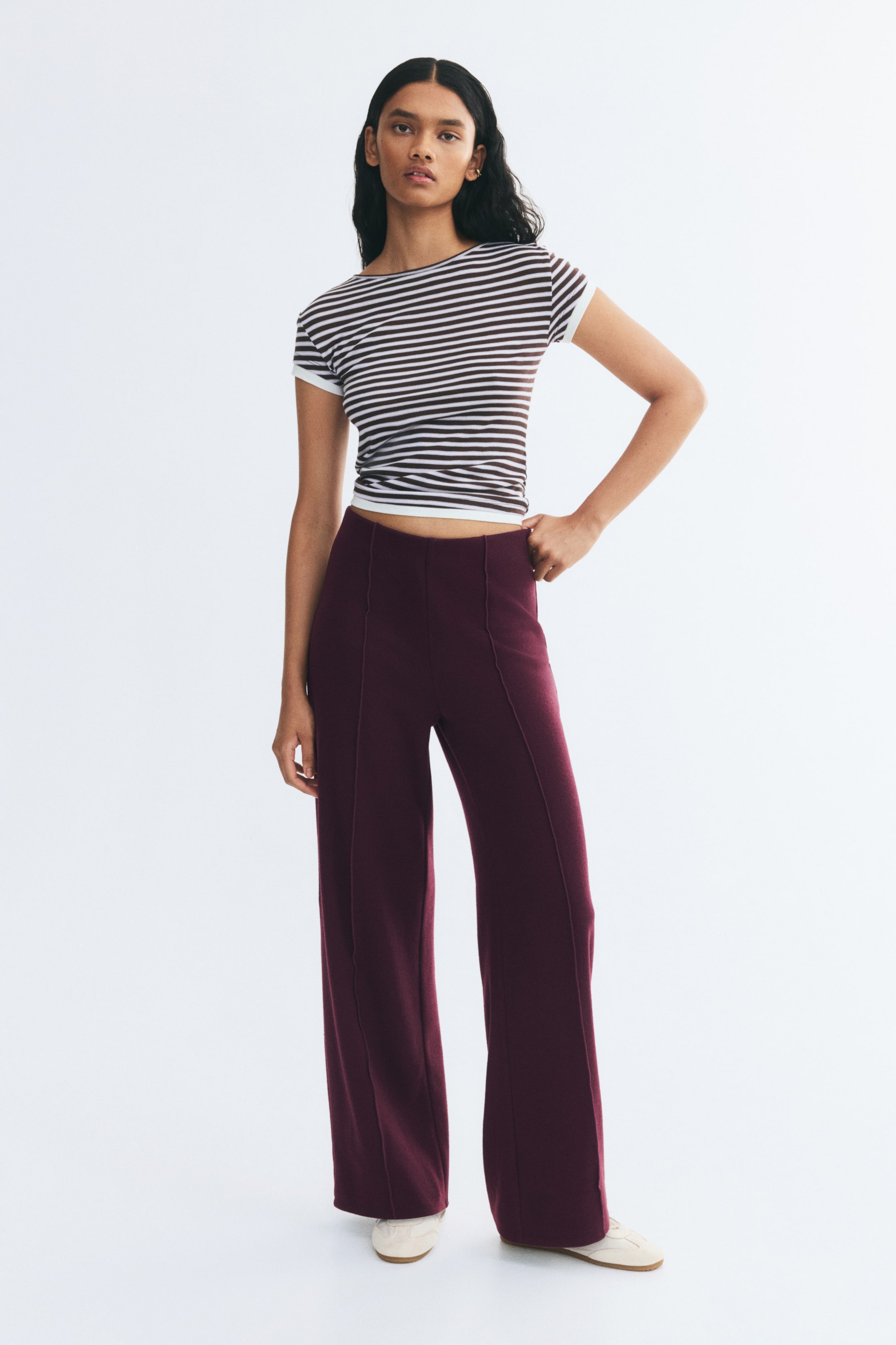 Ampliar la imagen: Pantalones de punto de corte holgado en color ciruela intenso, con cintura elástica y marcadas pinzas cosidas al frente de las piernas rectas, que lleva puestos la modelo. Los pantalones se combinan con una camiseta de manga corta a rayas y zapatos planos de color claro.