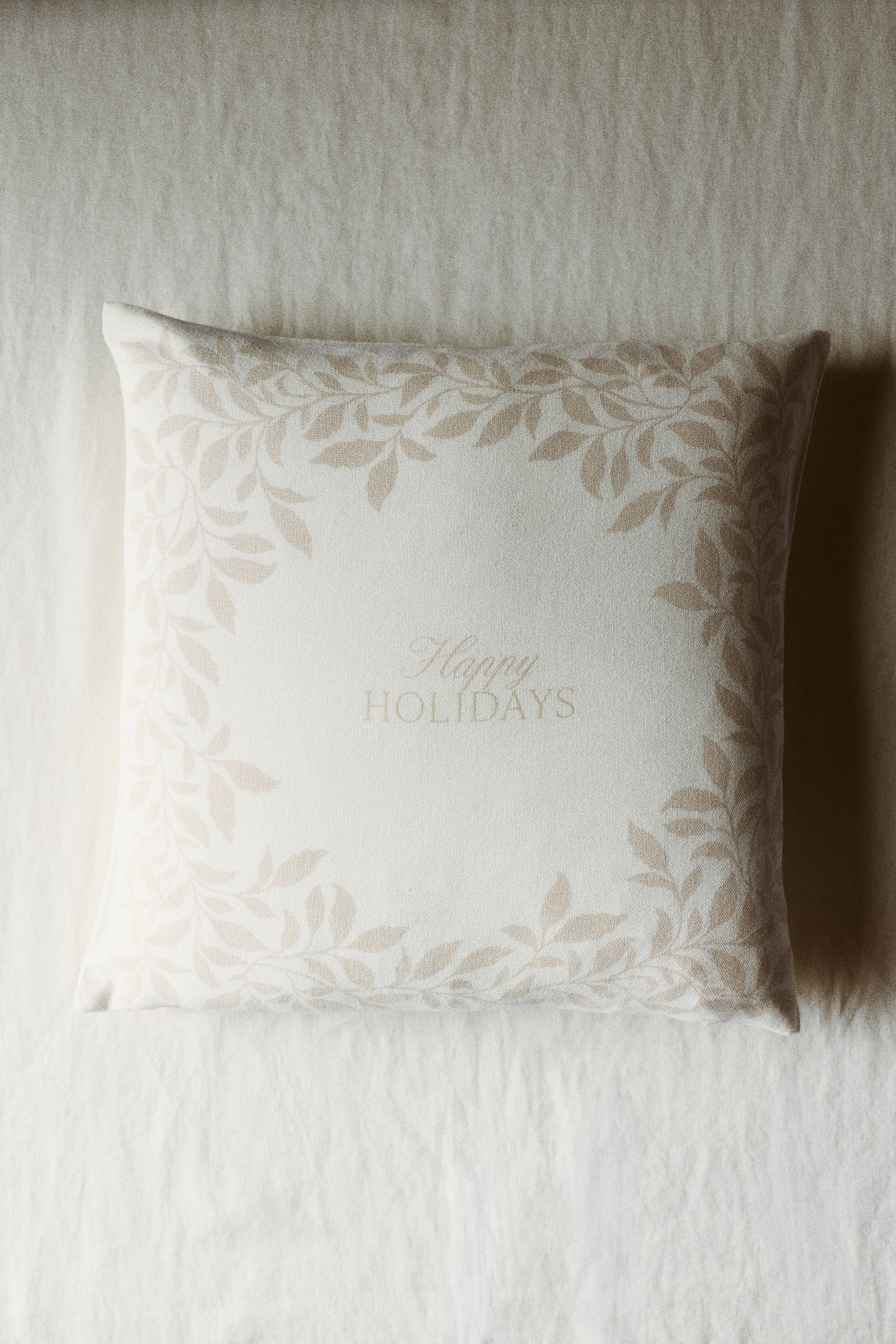 Et firkantet putetrekk i en off-white blanding av lin og bomull er vist forfra, med en trykt lys beige bladkant som rammer inn ytterkantene. Ordene Â«Happy HOLIDAYSÂ» er trykt i midten i en matchende lys beige skrift i manus- og serif-font.