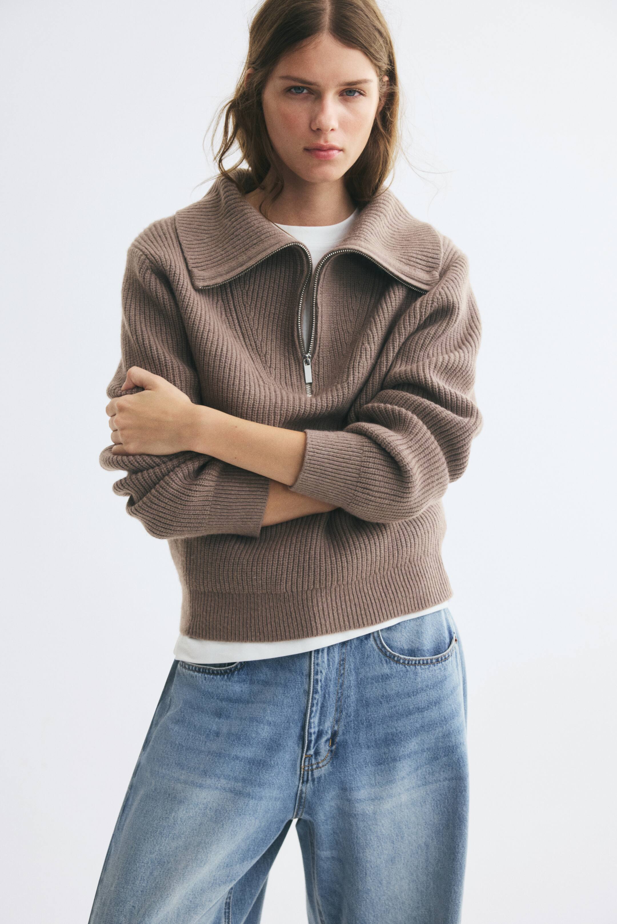 Visualizza l'immagine ingrandita: Un maglione in maglia a coste di colore marrone taupe è indossato dalla modella con le braccia incrociate, sopra una T-shirt bianca e jeans blu. Il maglione presenta un colletto con mezza zip lasciata aperta, maniche lunghe con polsini a coste e un orlo a coste.