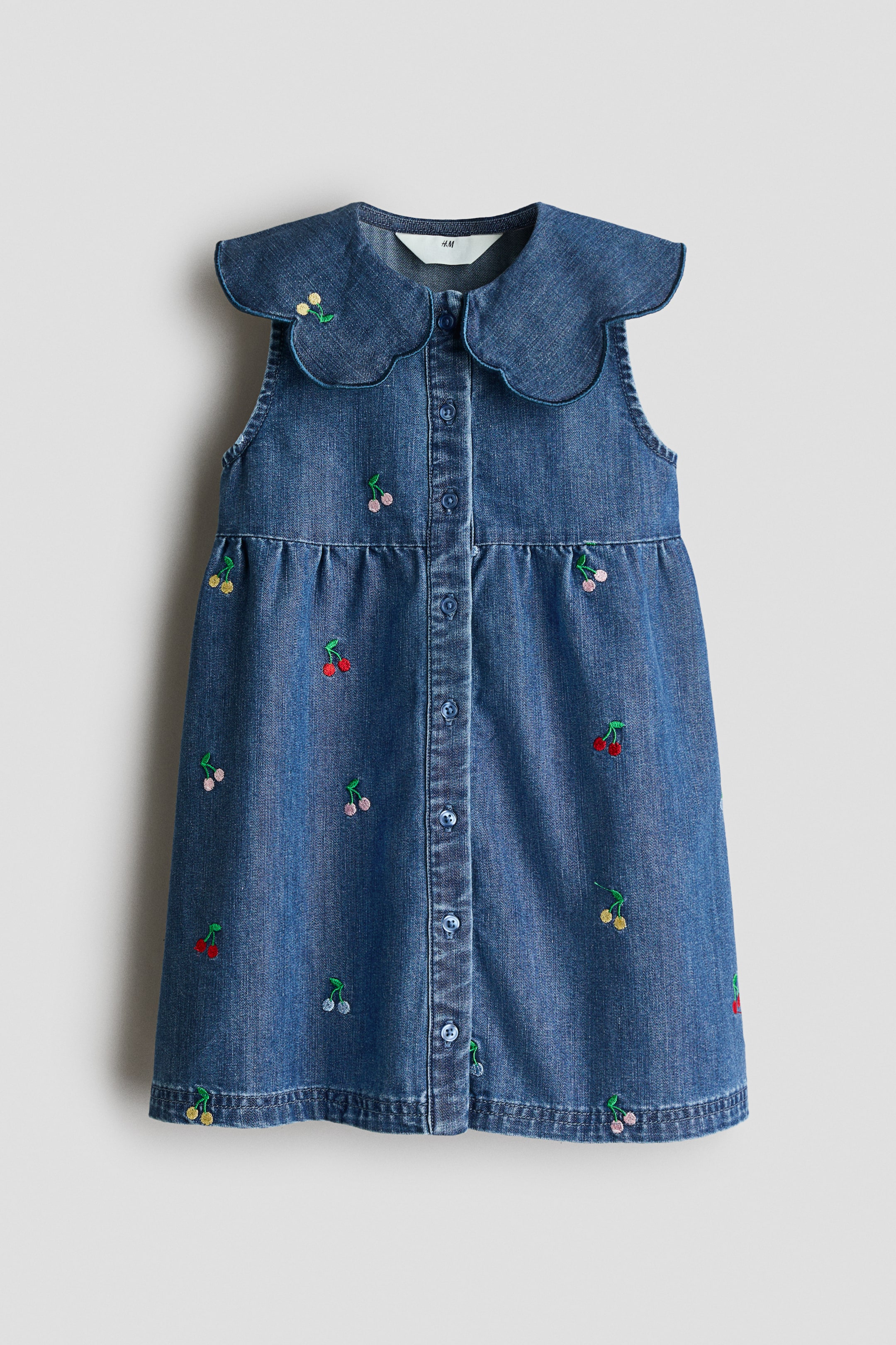 View larger image: A sleeveless blue denim dress is shown from the front, featuring all-over embroidered cherry motifs. A Peter Pan collar, edged with blue accent embroidery, frames the neckline, and buttons fasten down the front. A gathered seam defines the waist above a flared skirt.