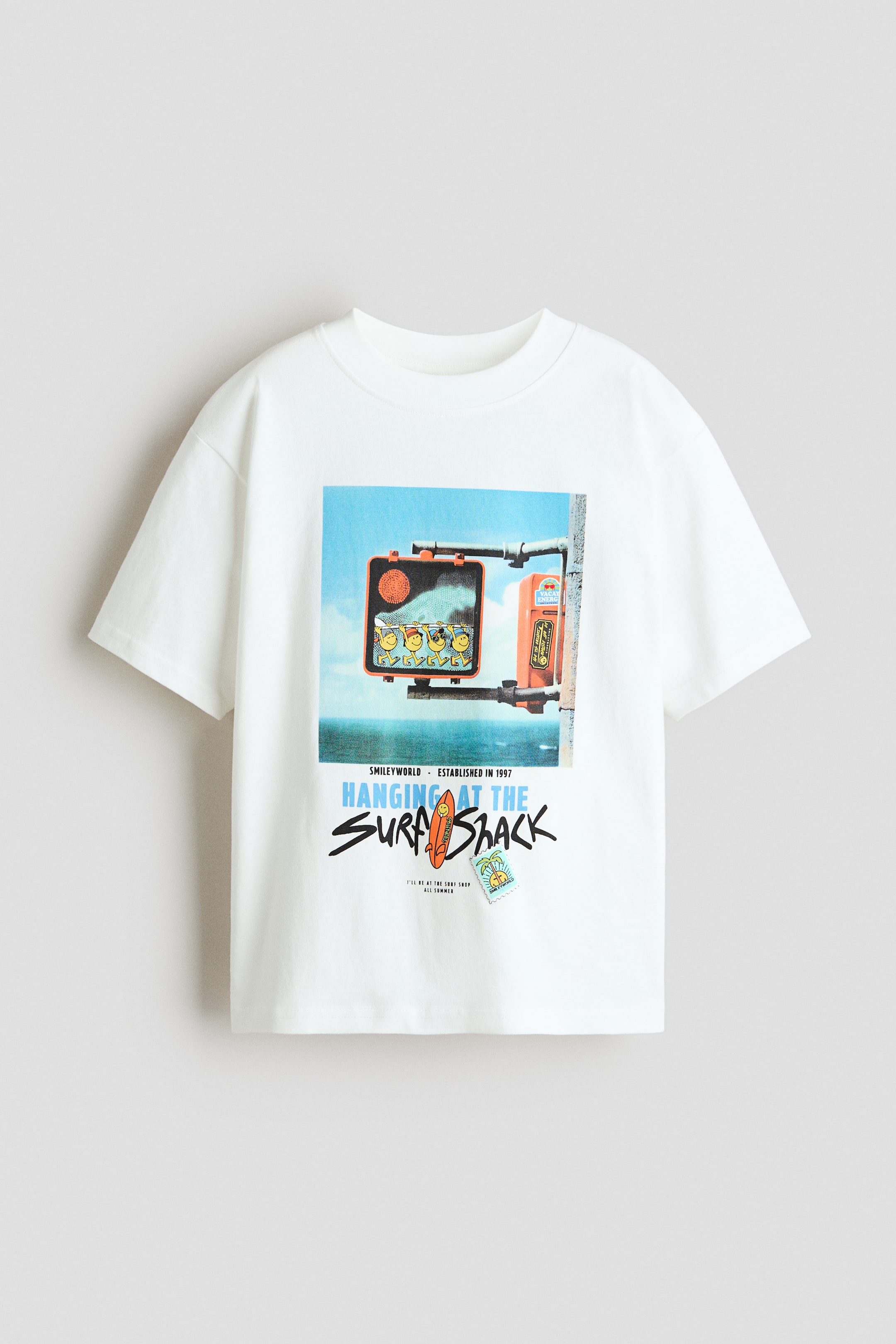 Grotere afbeelding bekijken: T-shirt van zachte katoenen jersey met een geribde afwerking rond de halslijn en een losse pasvorm met verlaagde schouders. De voorkant toont een kleurrijke grafische print van een straatscène met een verkeerslicht dat smiley gezichten weergeeft, de tekst 'hanging at the surf shack', een surfplank, en kleine stempels.