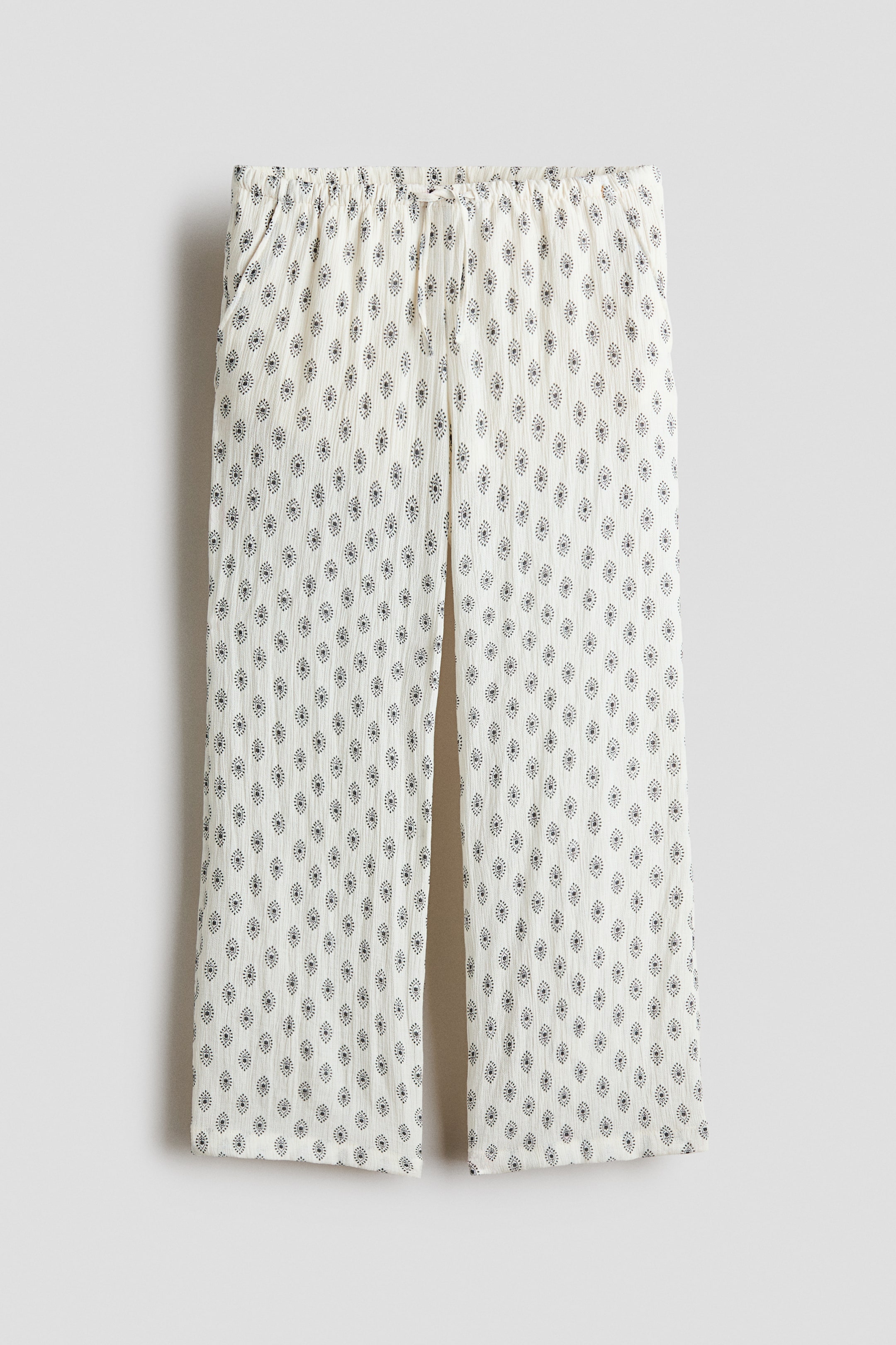 View larger image: Relaxed-fit trousers featuring a light-coloured woven fabric with a repeating dark diamond-shaped pattern. The front view showcases an elasticated, drawstring waist and diagonal side pockets.
