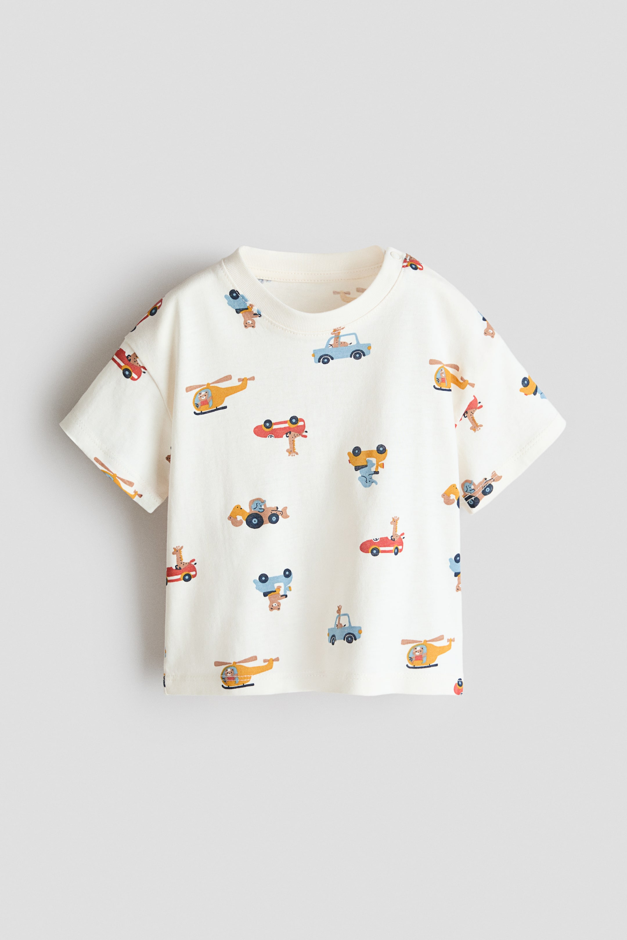 View larger image: A printed cotton T-shirt is displayed front-facing, in an off-white color. It features an all-over pattern of colorful vehicles with animals, a ribbed crew neck, short sleeves, and snap button closures on the left shoulder.