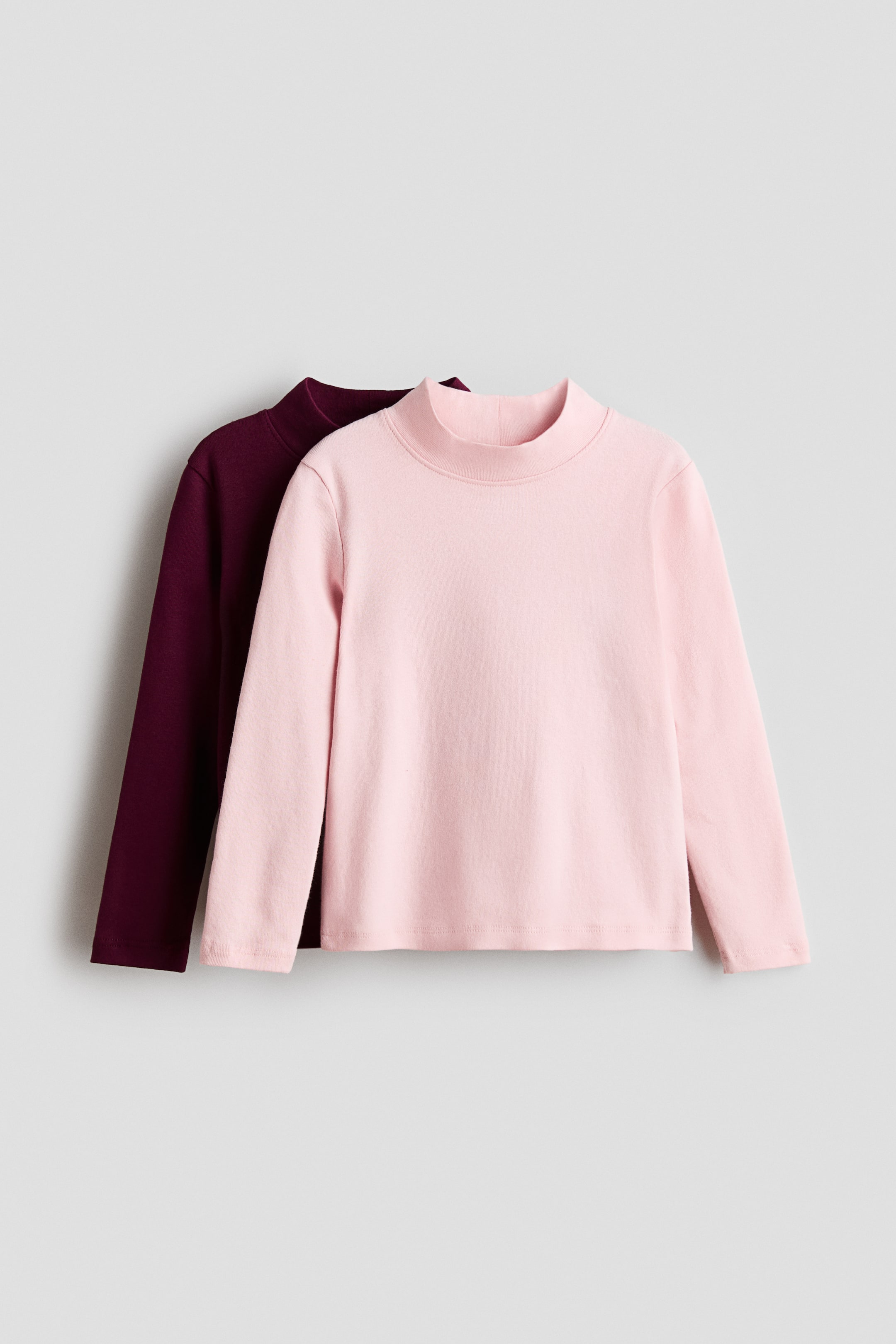 View larger image: Two long-sleeved mock neck tops are shown from the front, with a light pink top layered directly over a dark burgundy top. Both tops feature a high mock neckline, long sleeves, and a straight hem.