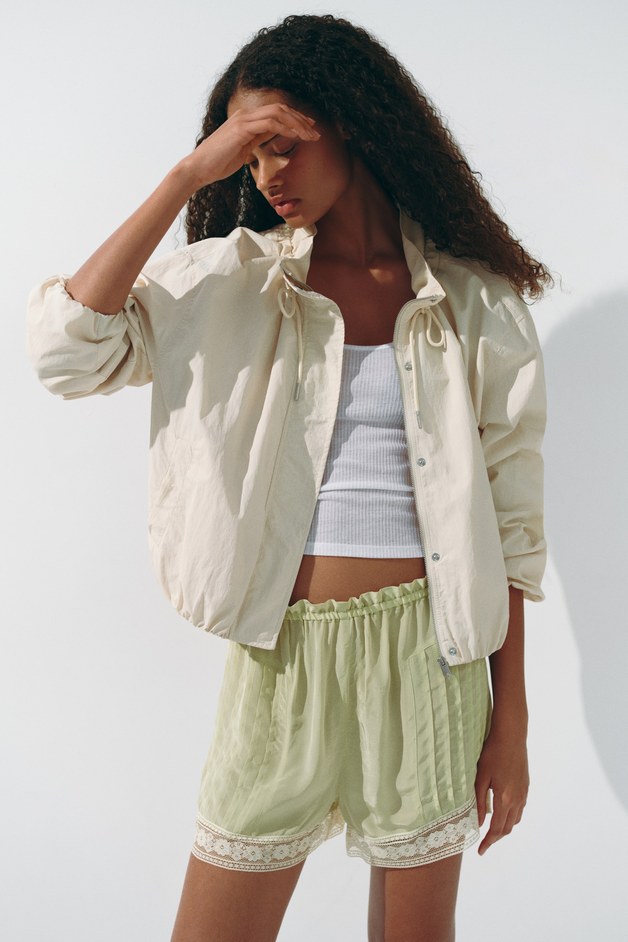 View larger image: A light beige lightweight jacket featuring a standing collar with drawstrings and a full zip closure, partly concealed by a snap placket. The jacket showcases voluminous long sleeves with elasticated cuffs and an elasticated hem, worn open over a white ribbed vest top.