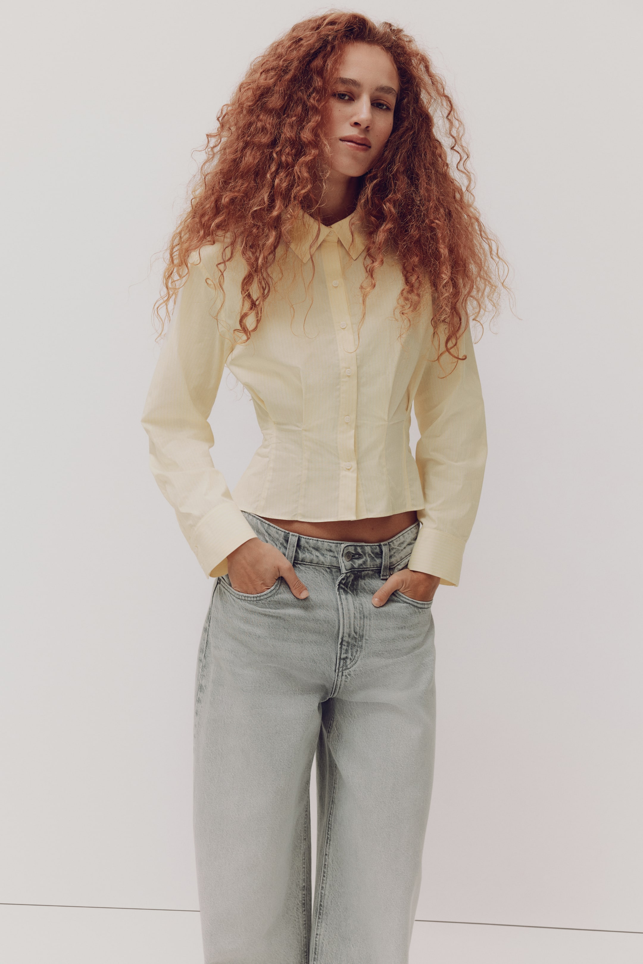 Xem ảnh lớn hơn: Chiếc áo sơ mi cài khuy màu vàng nhạt, ôm vừa vặn, có cổ bẻ kiểu cổ điển, tay dài với cổ tay cài nút, eo được thắt nhẹ và vạt áo cắt ngắn. Áo được phối cùng quần jean wash nhạt, cạp cao, dáng ống rộng thoải mái.