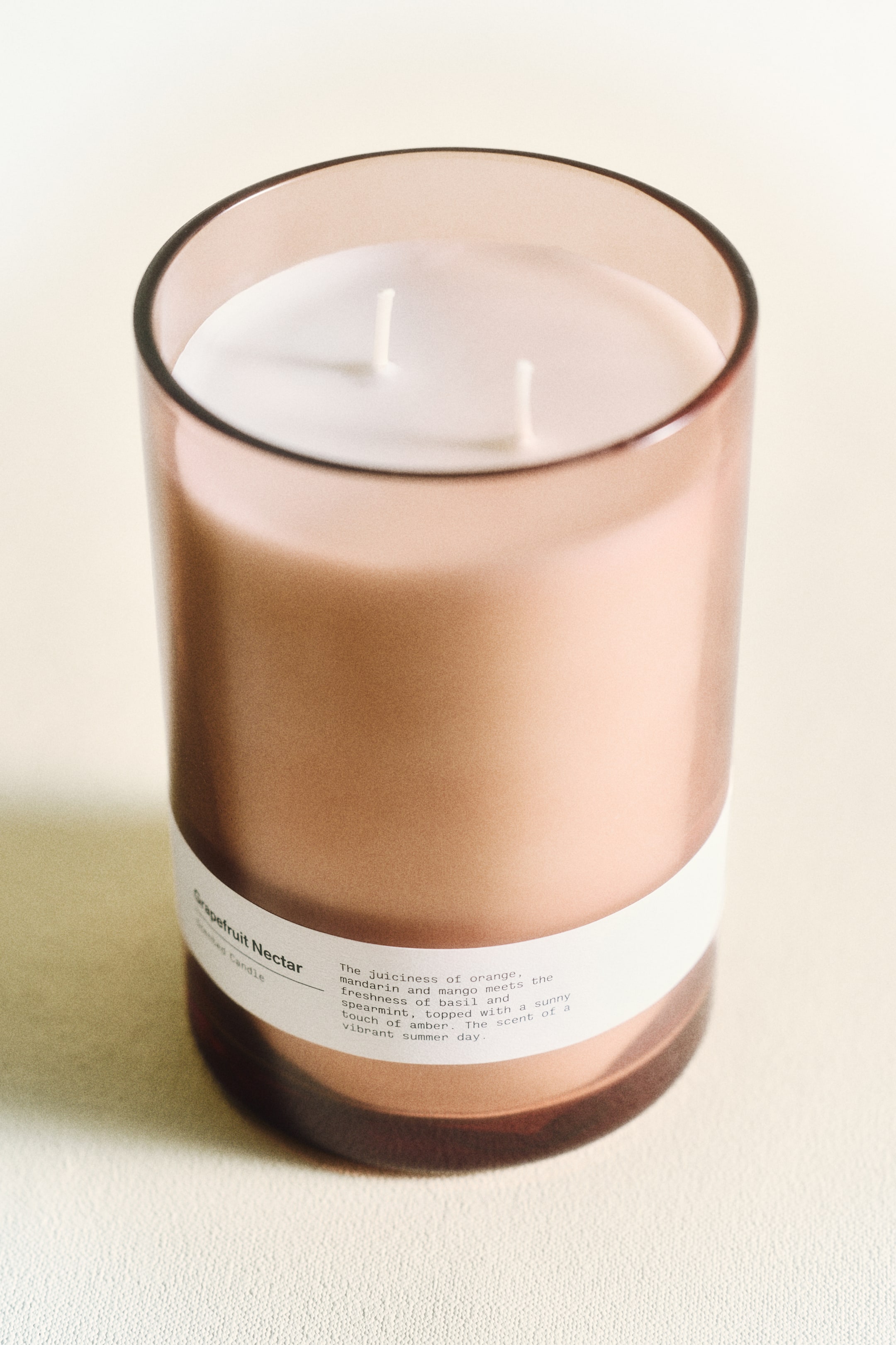 הצגת תמונה גדולה יותר: נר מוצג במיכל זכוכית גלילי, שקוף בצבע חום-ורוד, הנצפה מזווית מעט מוגבהת. שני פתילים לבנים עומדים זקופים בשעווה הלבנה, ותווית לבנה עוטפת את החלק התחתון של המיכל, מציגה את הכיתוב "נקטר אשכוליות" ותיאור הריח.