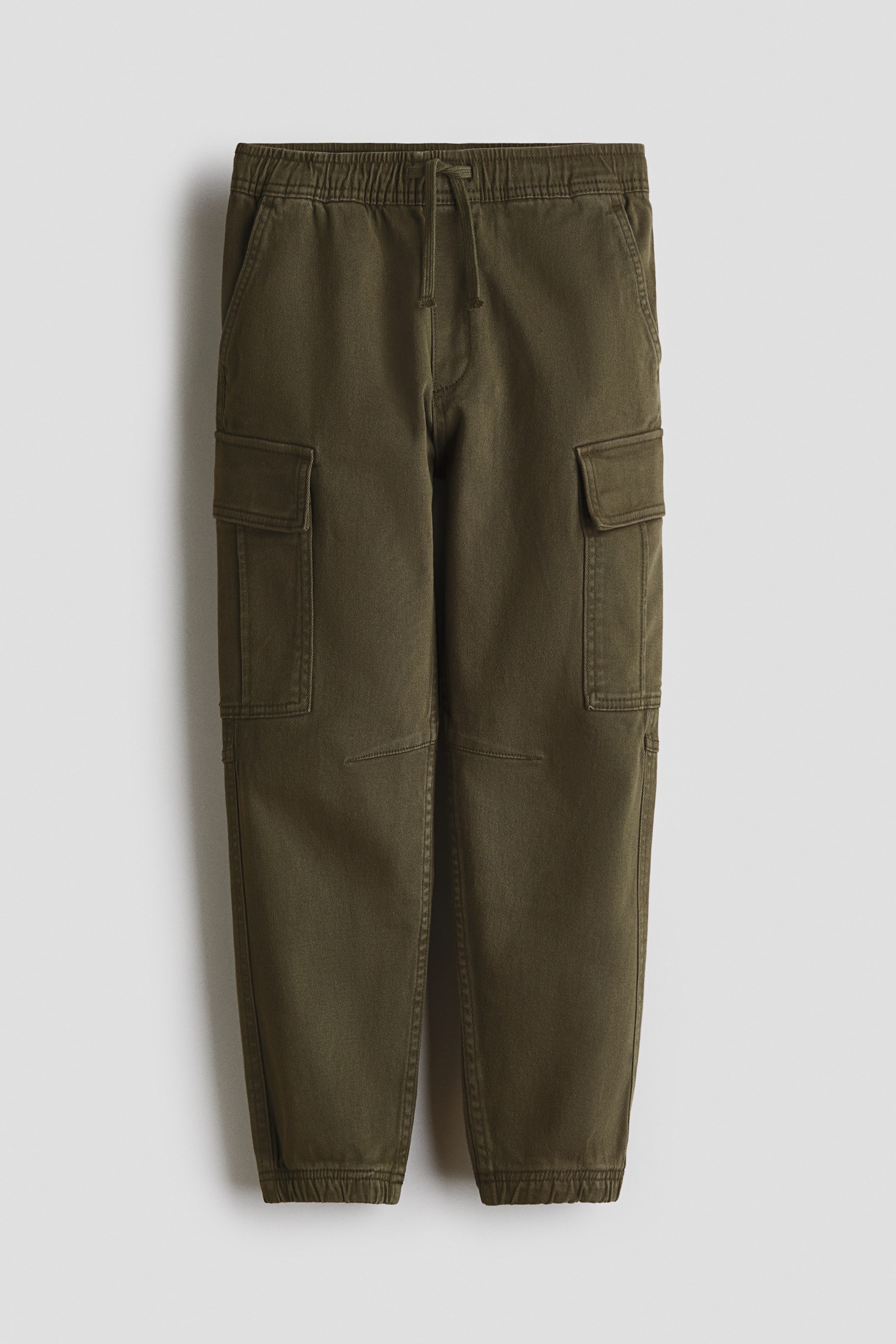 View larger image: Olive green cotton twill cargo joggers are shown from the front, highlighting an elastic drawstring waist, a visible faux fly, and front slash pockets. The legs feature flap cargo pockets on the sides and covered elastic at the hems.