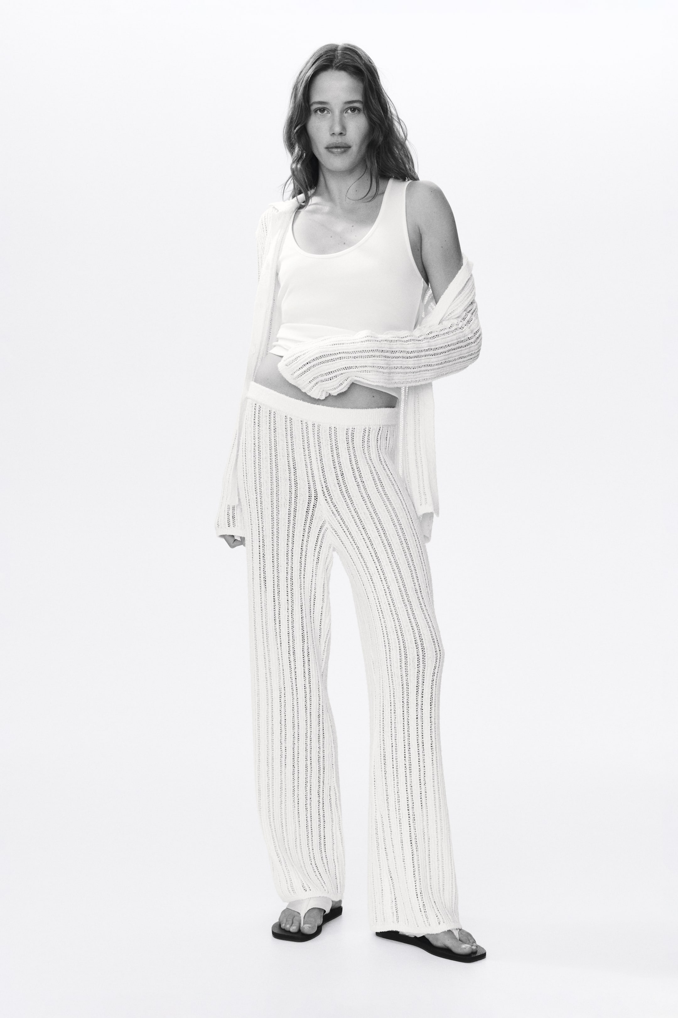 Agrandir l'image: Un cardigan blanc à manches longues en maille ajourée et un pantalon large assorti sont présentés. Le cardigan transparent à rayures verticales est drapé sur un débardeur blanc à encolure dégagée, avec ses longues manches partiellement portées, et le pantalon taille haute s'étend jusqu'au sol.