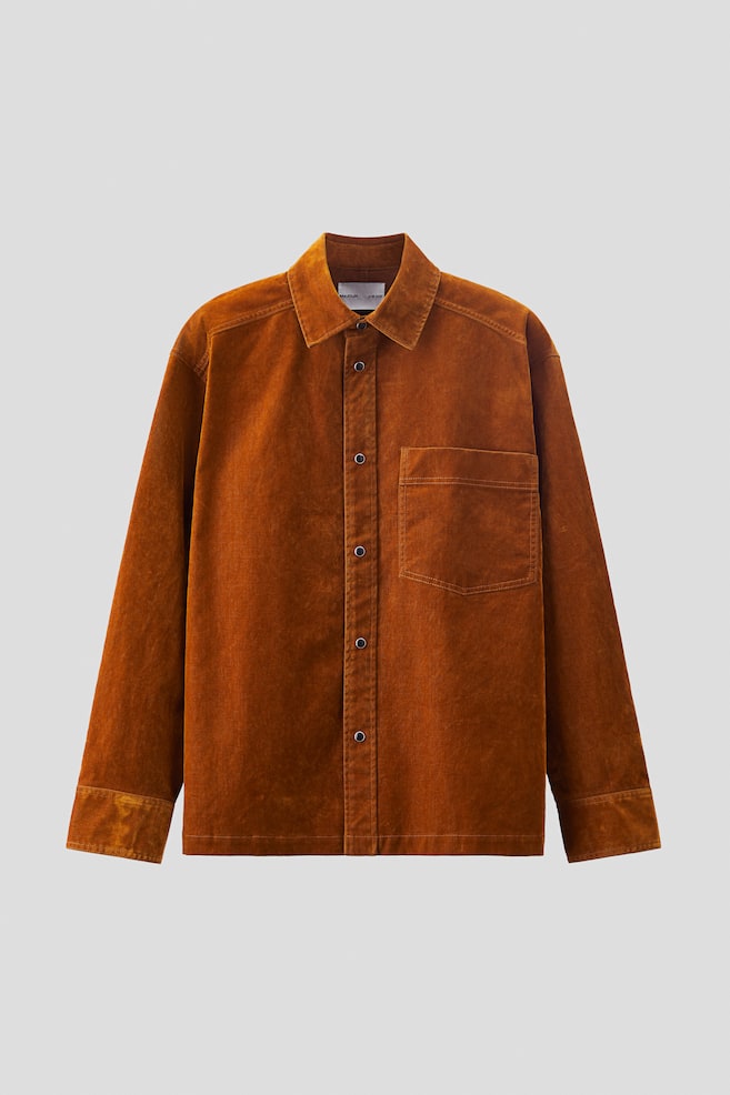 Une surchemise affiche une coupe ample dans un tissu brun rouille chaud et brossé, tournée vers l'avant. Elle est dotée d'un col classique, d'un devant avec des boutons-pression sombres, de manches longues avec des poignets droits, d'une poche poitrine unique sur la gauche et d'un ourlet droit. - 1