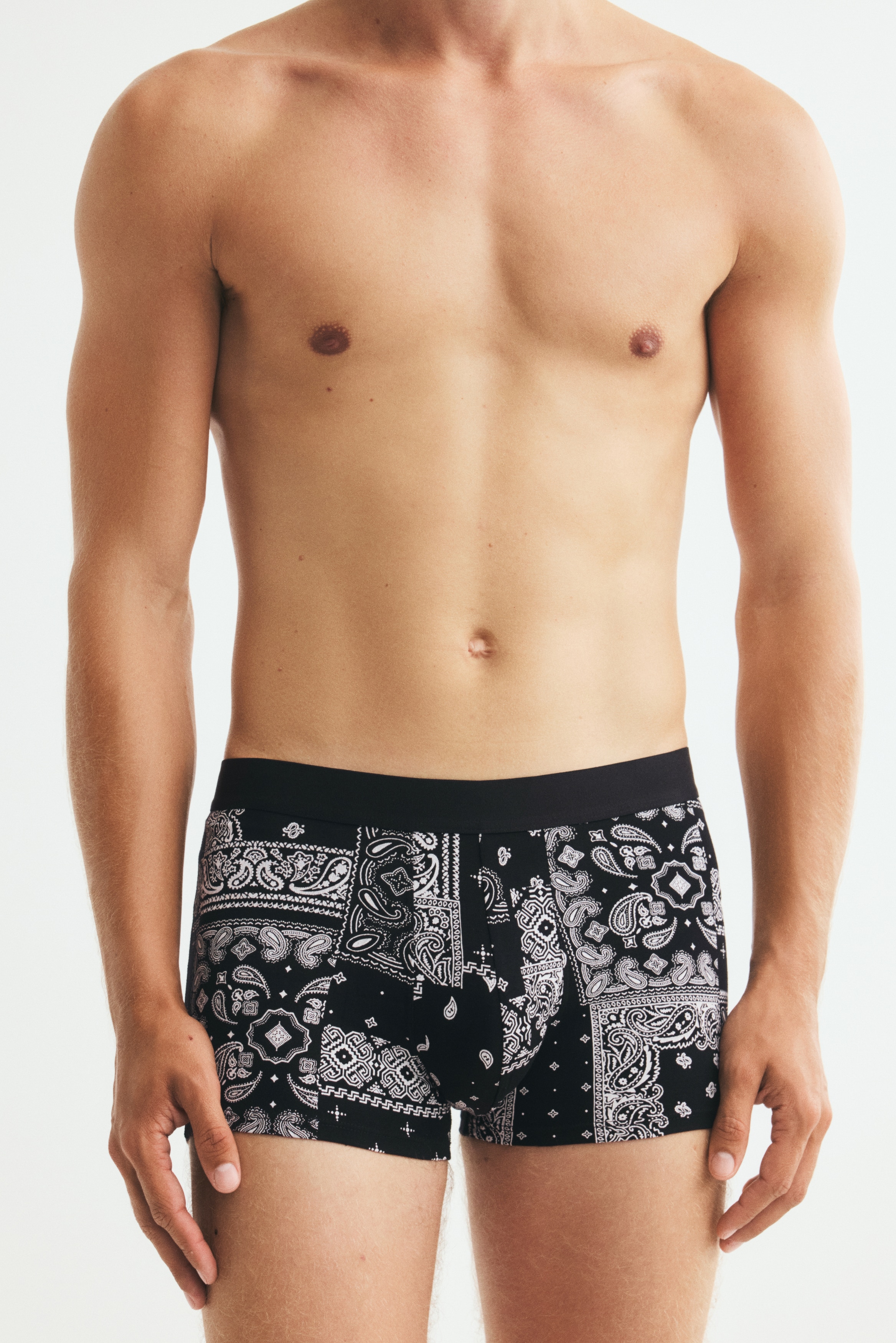 Drei Paar Boxershorts präsentieren ihre Vorderseiten, gestapelt mit sichtbaren Bündchen. Das vorderste Paar zeigt ein schwarz-weißes Bandana-Muster, während das mittlere Paar dunkelgrau ist und das oberste Paar dunkelrot, beide mit dezenten tonalen Bandana-Mustern. Alle verfügen über verdeckte, elastische Bündchen und kurze Beine.