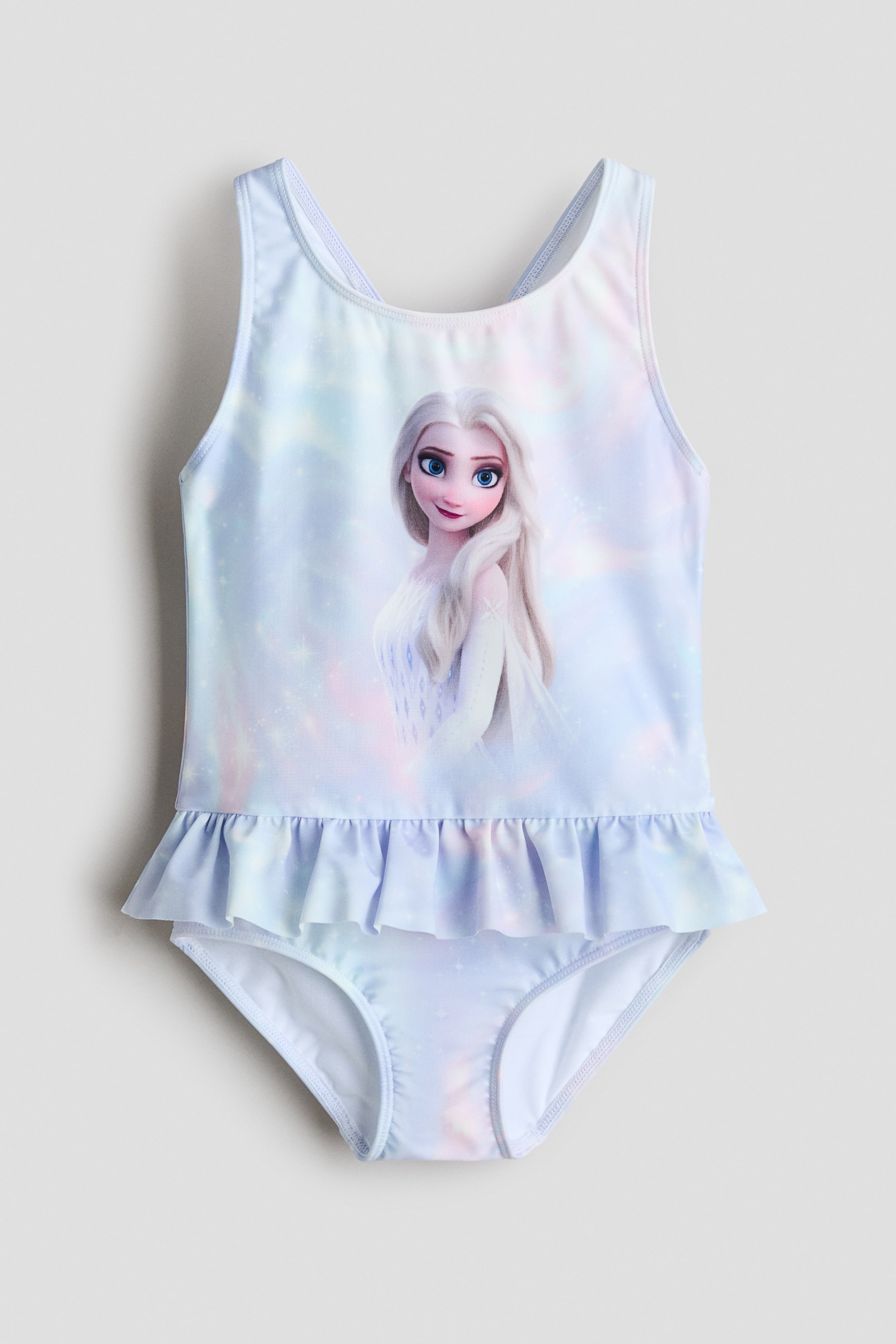 Visa större bild: En barnbaddräkt i helmodell med ett ljusblått, rosa och vitt akvarellliknande mönster, med ett grafiskt tryck av Elsa från Frozen på framstycket och en volangdetalj i form av en kjol runt midjan.