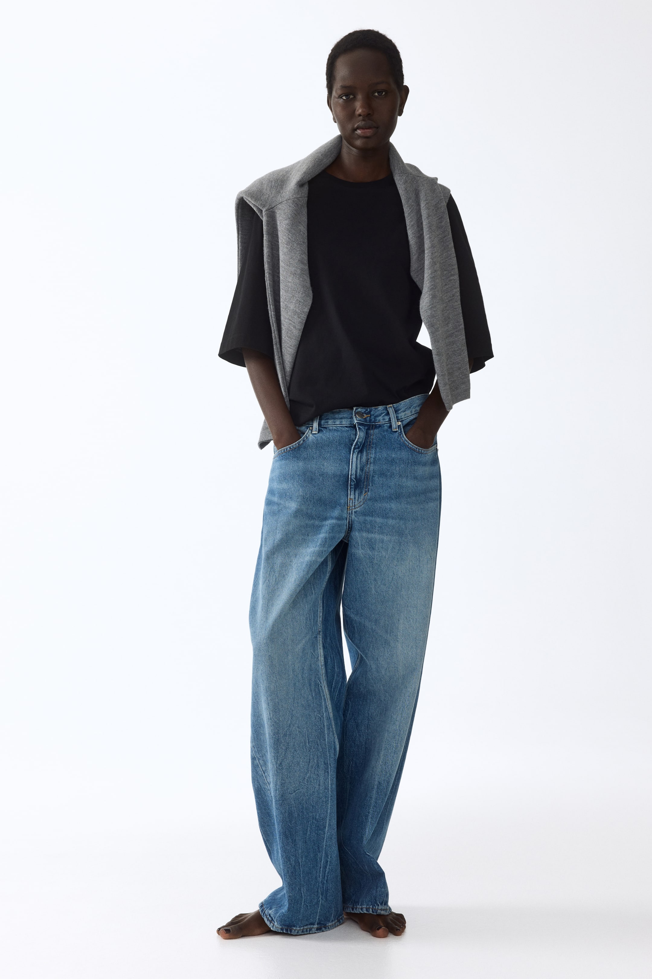 查看大圖: 一件黑色圓領短袖T恤，寬鬆的短袖設計，搭配一條高腰藍色寬腿牛仔褲，牛仔褲呈現中等水洗效果，褲腳堆疊在腳踝處。一件灰色針織毛衣披在肩上，搭在T恤外。