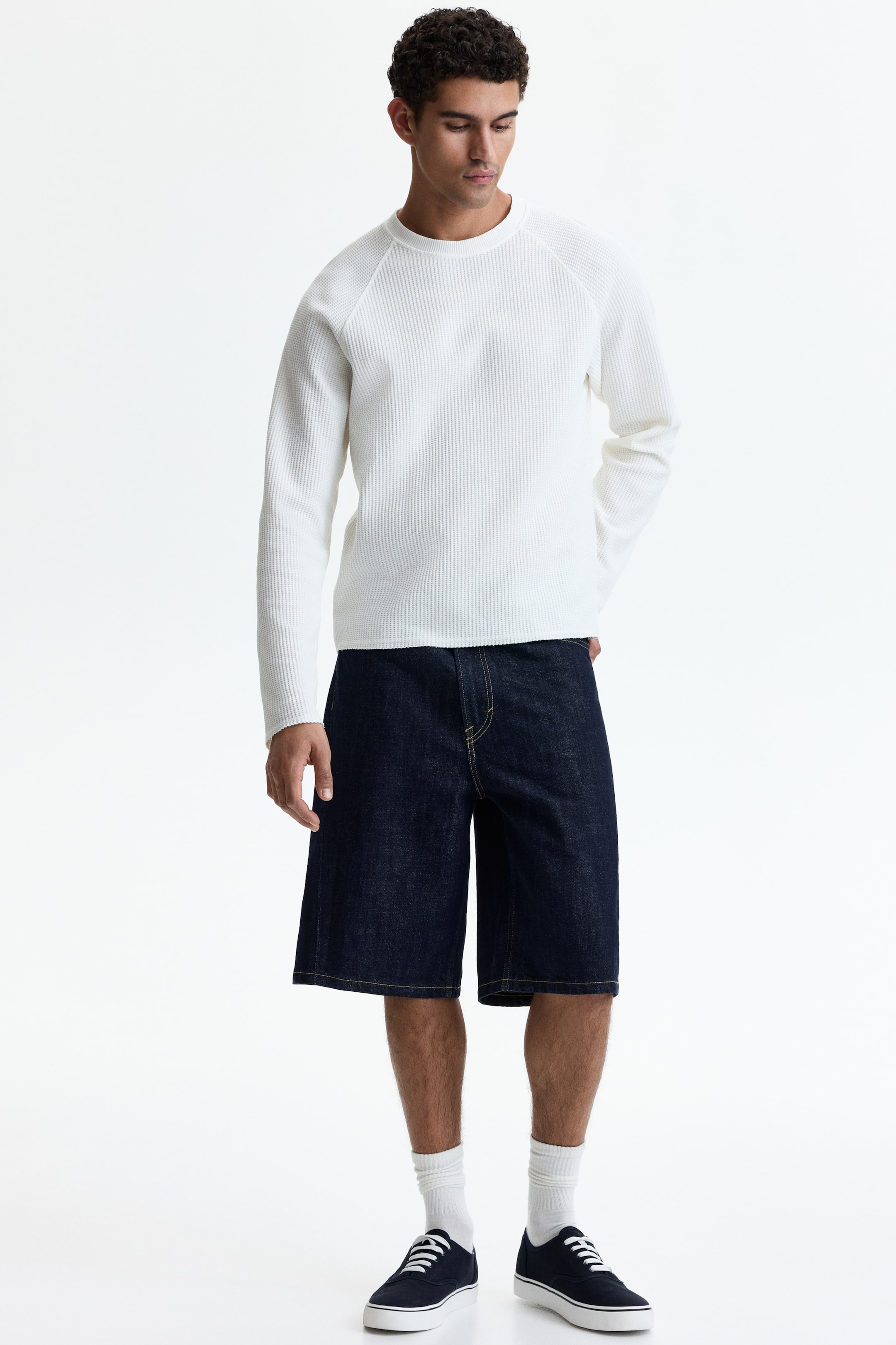 View larger image: 5-pocket shorts in dark wash rigid cotton denim, featuring forward-facing side seams, a rounded leg, and a baggy fit from the seat to the hem with a dropped crotch, regular waist, and zip fly and button, are being worn by the model. The model is styling the shorts with a white long-sleeve top and dark sneakers.
