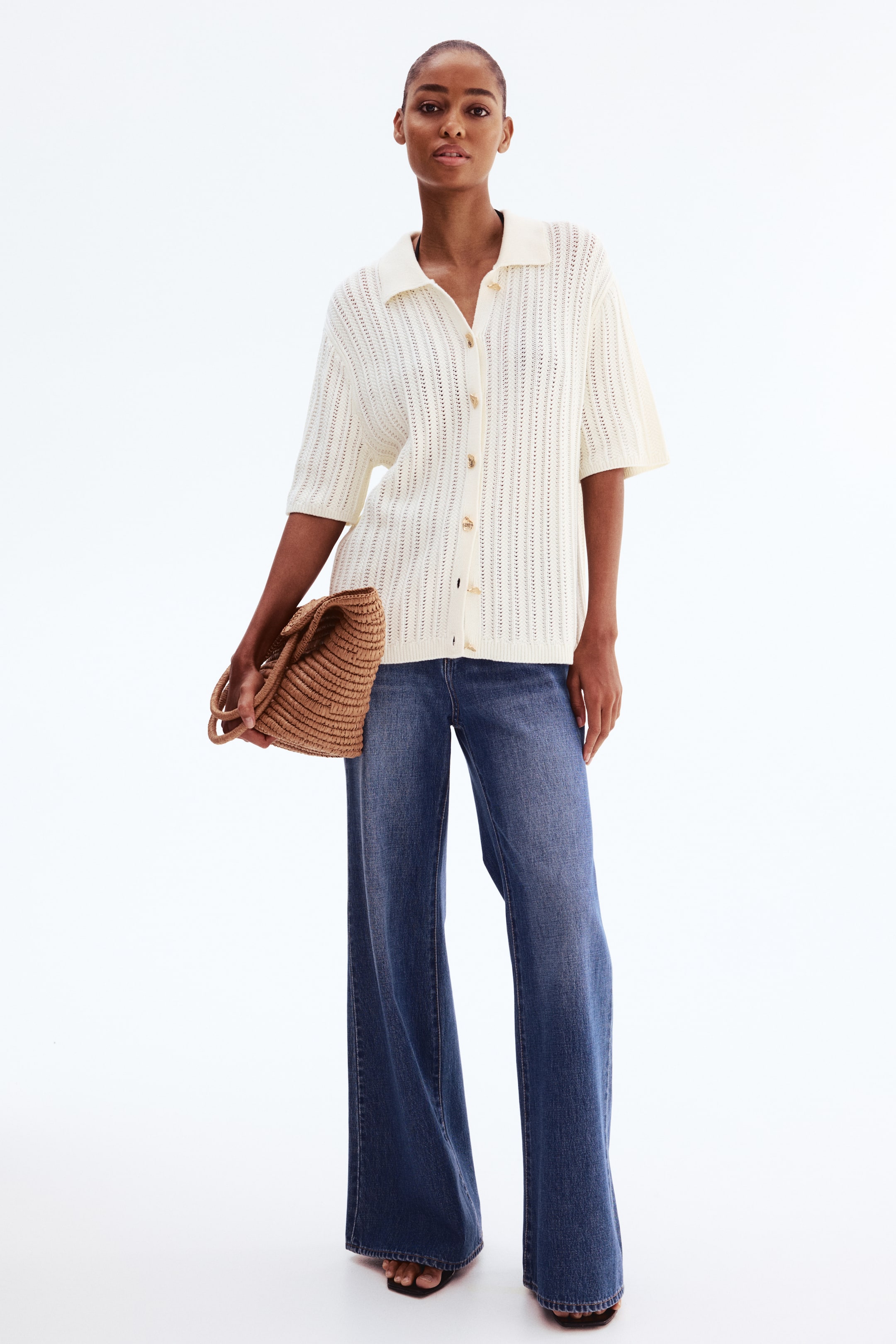 Ampliar la imagen: La modelo lleva puesto un cárdigan color crema en punto de algodón suave con apariencia de crochet. Presenta cuello, botones metálicos texturizados a lo largo del frente, hombros caídos, mangas hasta el codo y canalé en los puños y el dobladillo.