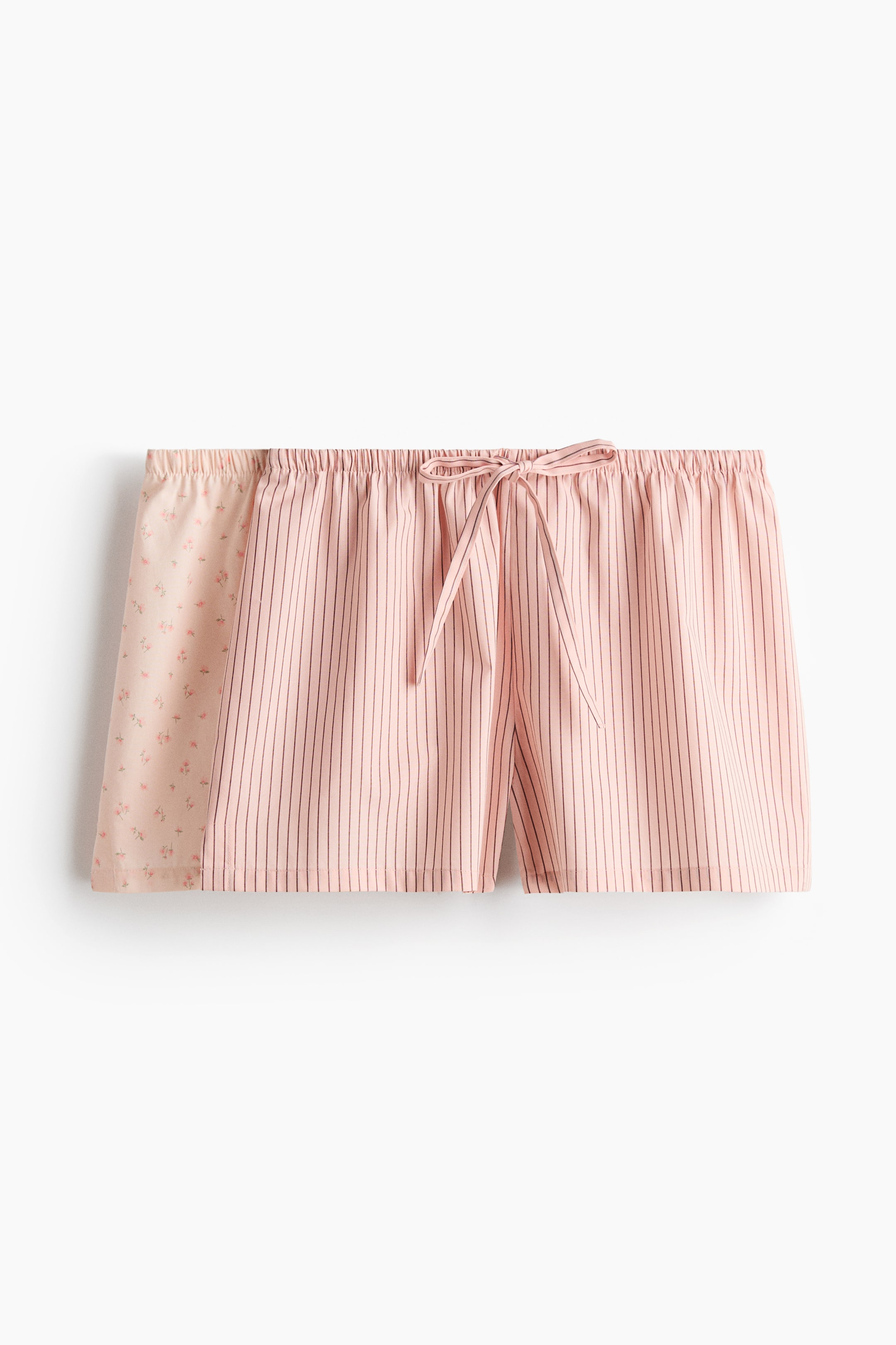 Se større billede: Pyjamashorts i vævet stof præsenterer et todelt design med et lyserødt blomstermønster på venstre side og lyserøde lodrette striber på højre side. En elastisk talje med snøre er synlig øverst, bundet i en sløjfe, med små slidser ved sømmene.