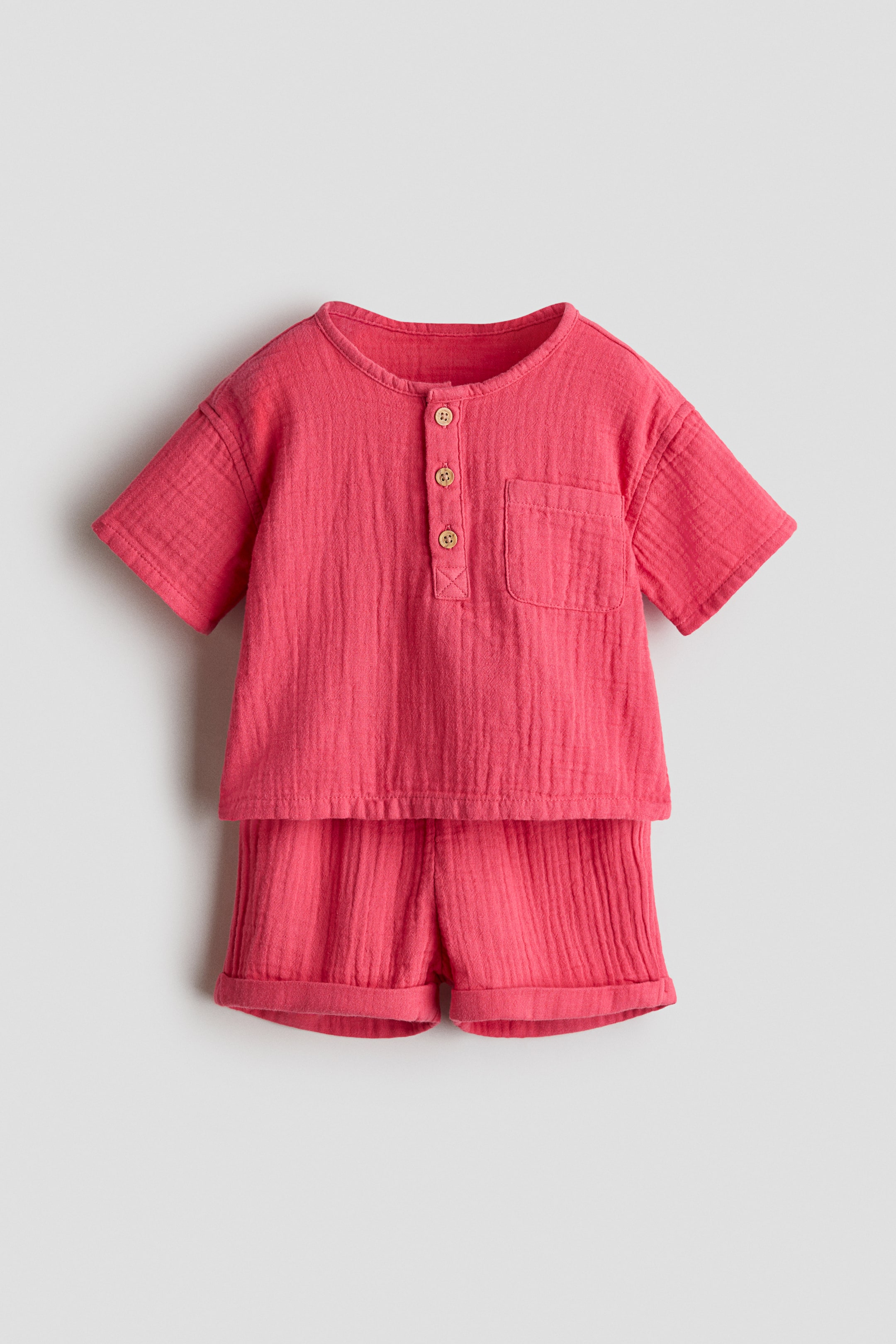 View larger image: Presenting a pink soft cotton muslin set, featuring a short-sleeved top with a round neckline, button placket, patch chest pocket, and dropped shoulders. Paired with matching shorts that have sewn-in turn-ups at the hems.