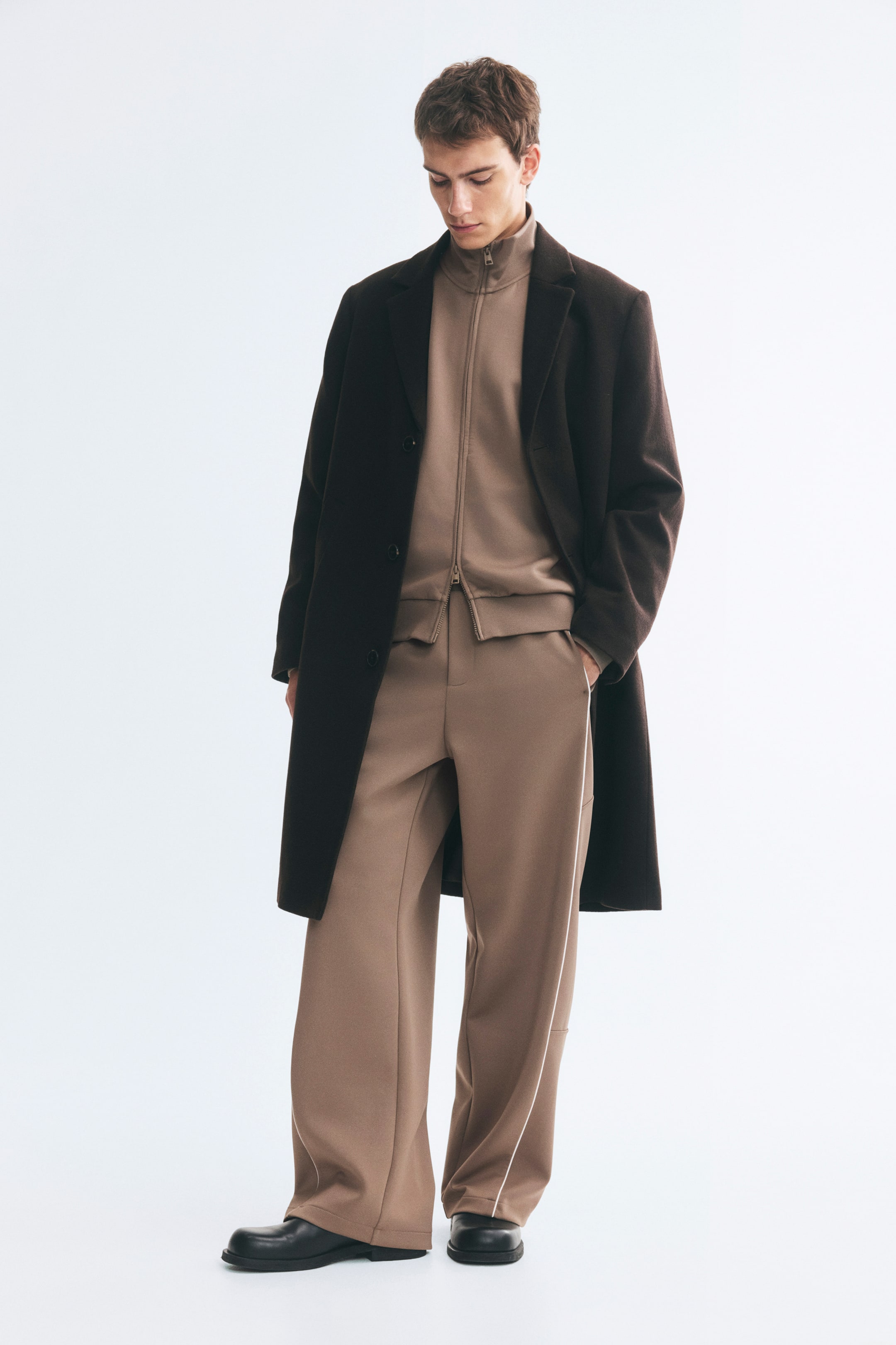 Größeres Bild ansehen: Taupefarbene Trackpants aus glattem Stoff werden vom Model getragen und zeichnen sich durch eine lockere Passform, gerade Beine und feine weiße Paspeln an den Seiten aus. Das Model steht mit einer Hand in der Tasche und kombiniert die Hose mit einer passenden Zip-up-Jacke, einem langen dunkelbraunen Mantel und schwarzen Schuhen.