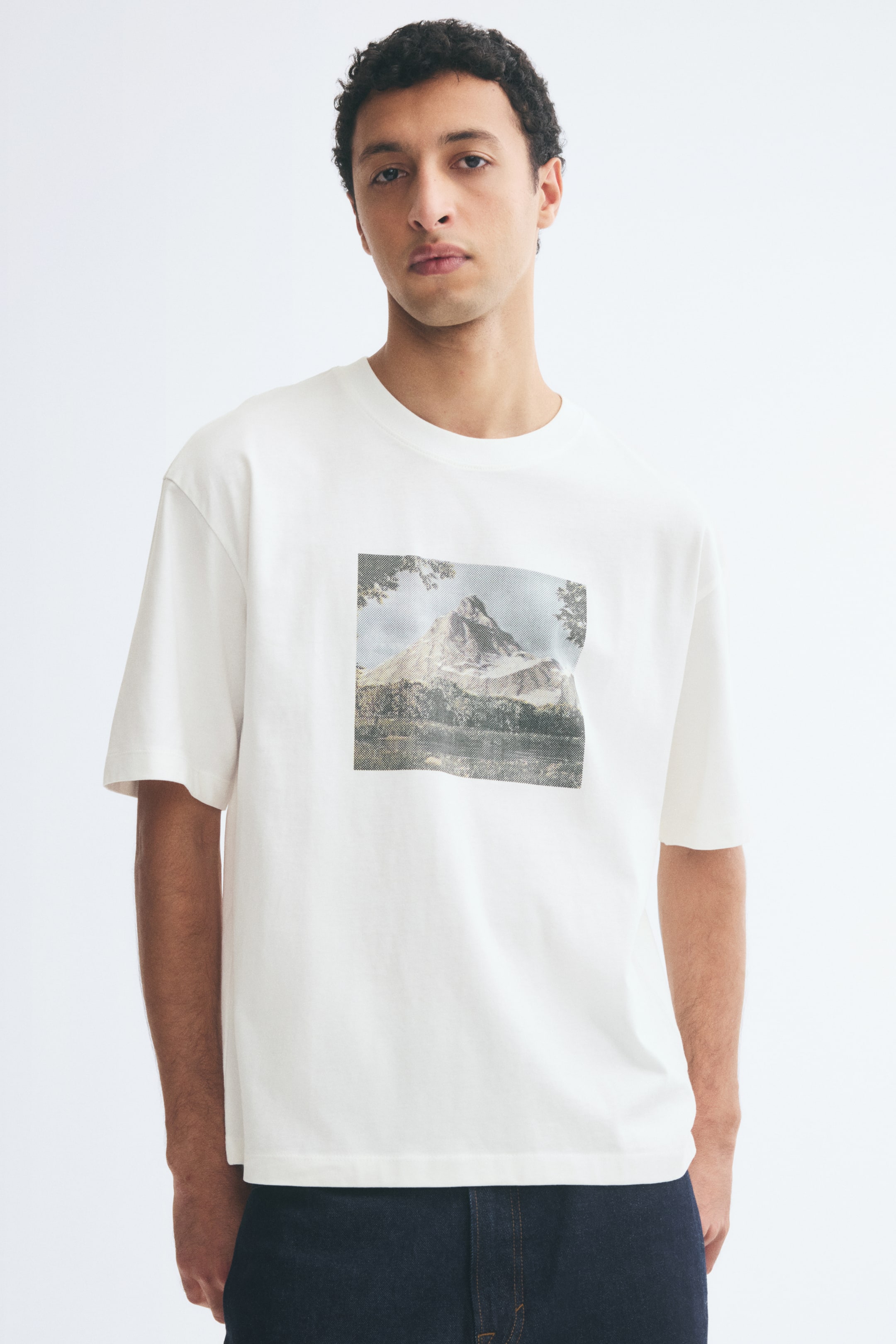 View larger image: The model is wearing an off-white midweight jersey T-shirt featuring a square graphic print of a mountain landscape. The relaxed-fit tee features a round ribbed neckline, dropped shoulders, and a straight hem, and is paired with dark blue jeans.