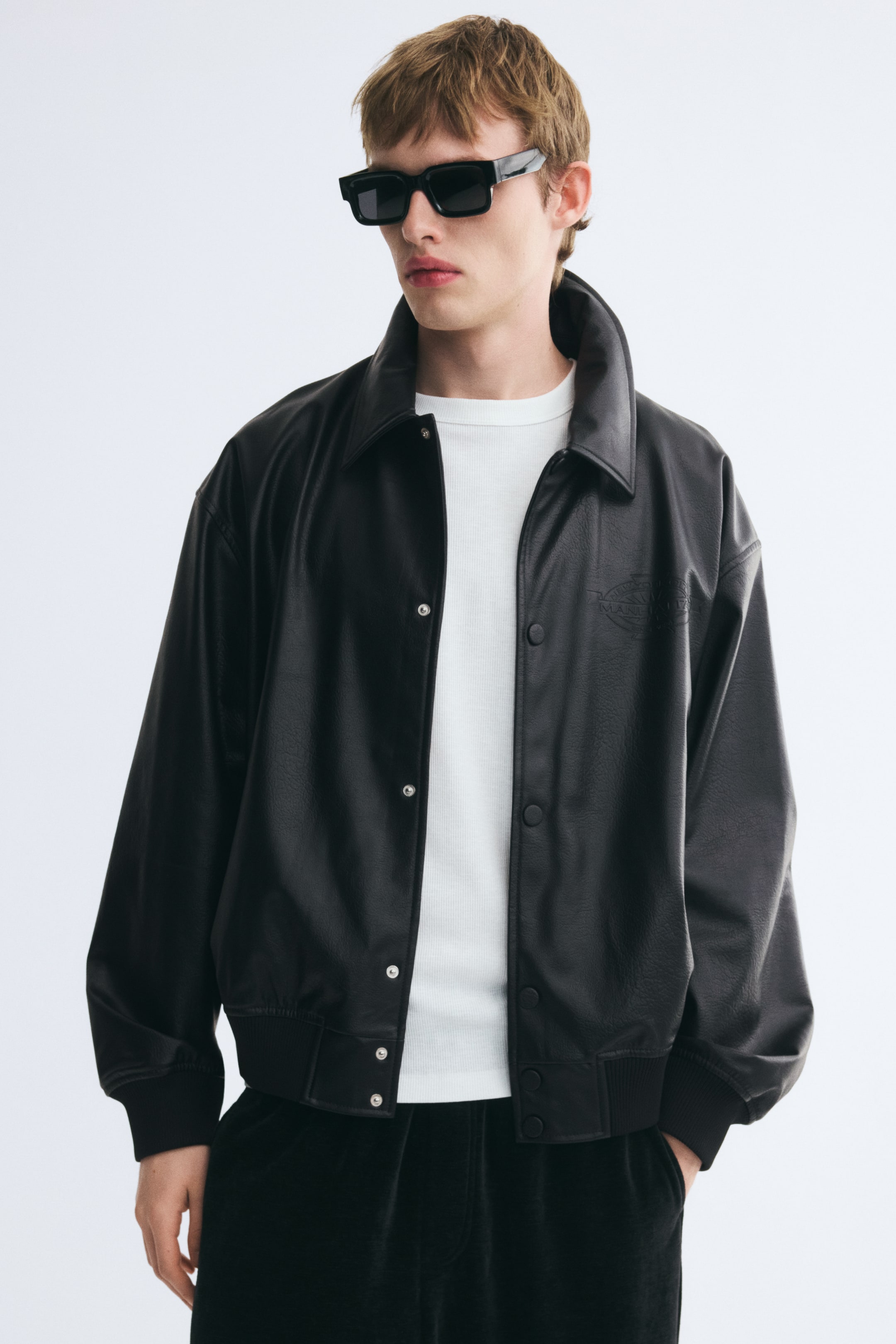 View larger image: A black jacket in coated fabric is being worn open by the model over a white T-shirt. The jacket features a collar, visible press studs down the front, and wide, ribbed trim at the cuffs and hem.