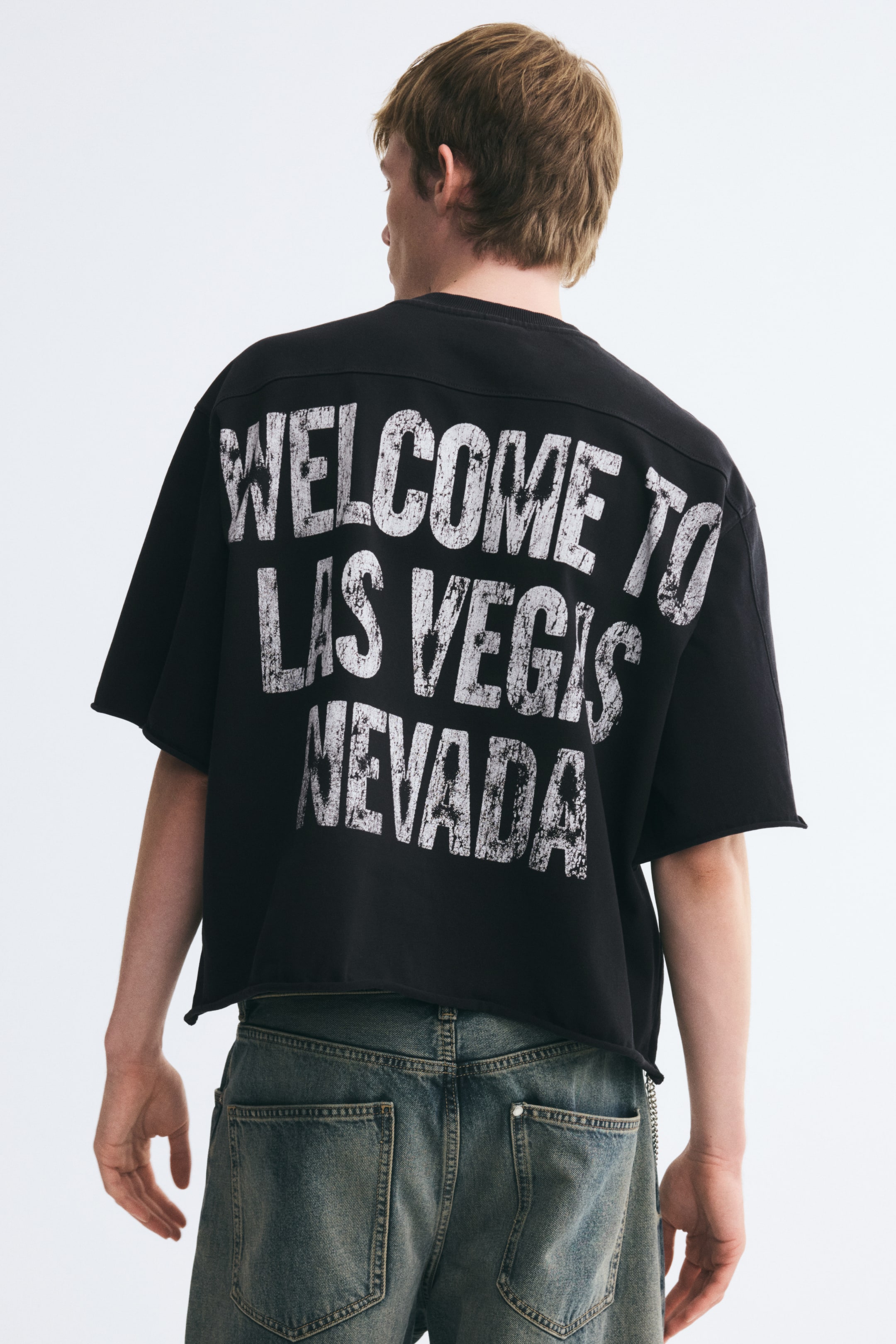 View larger image: A black, boxy, oversized T-shirt with dropped shoulders and a raw-cut hem is being worn by the model. The shirt features a large, distressed white graphic print across the back and hangs loosely over blue denim jeans.