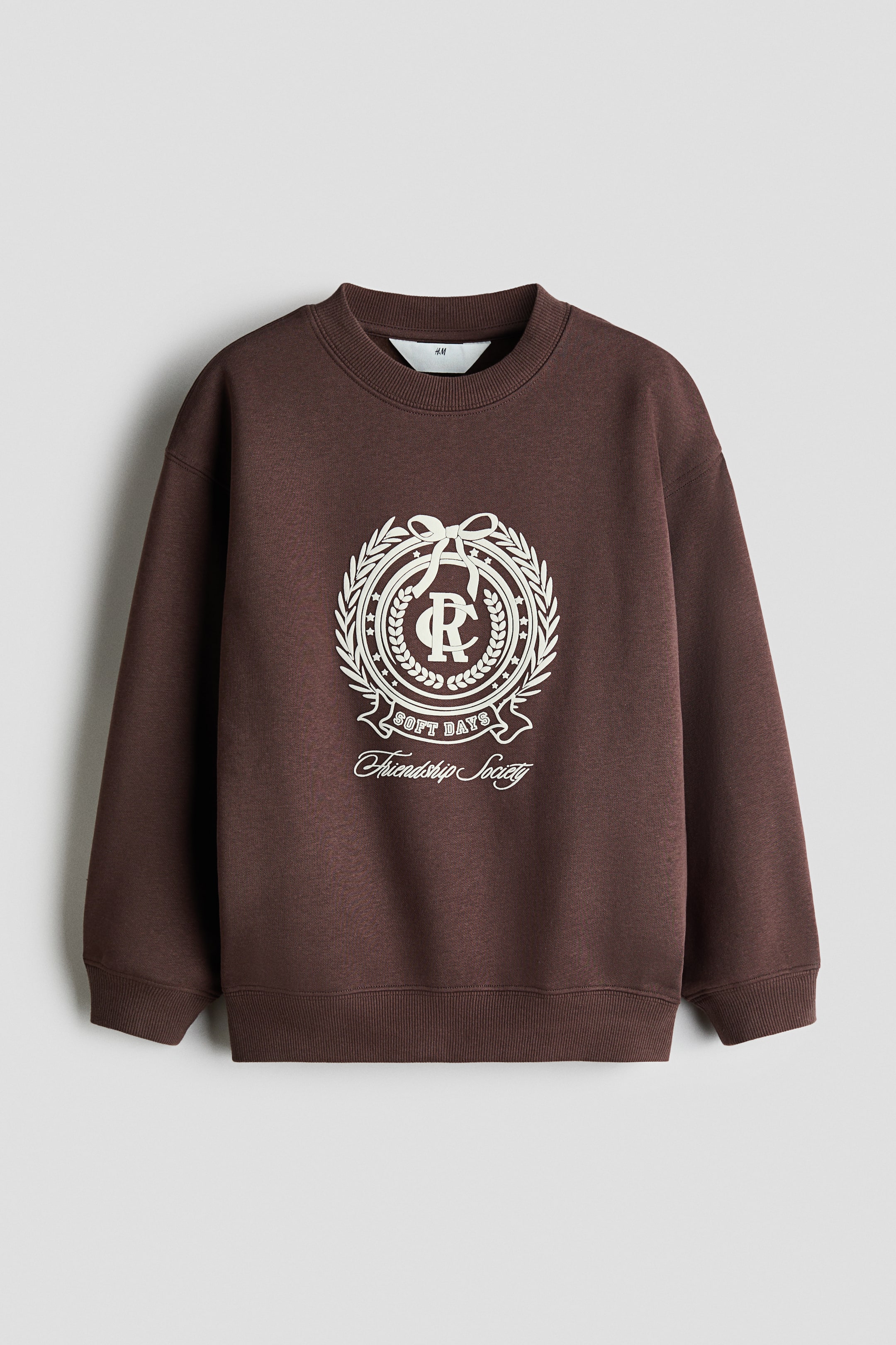 Vis større bilde: En mørk brun oversized sweatshirt vises forfra, med rund hals med ribbestrikket kant, droppede skuldre og bred ribbestrikket kant ved ermer og nederkant. Foran er det en kremfarget crest-grafikk med en sentral monogram, en laurbærkrans og teksten «Soft Days» og «Friendship Society».
