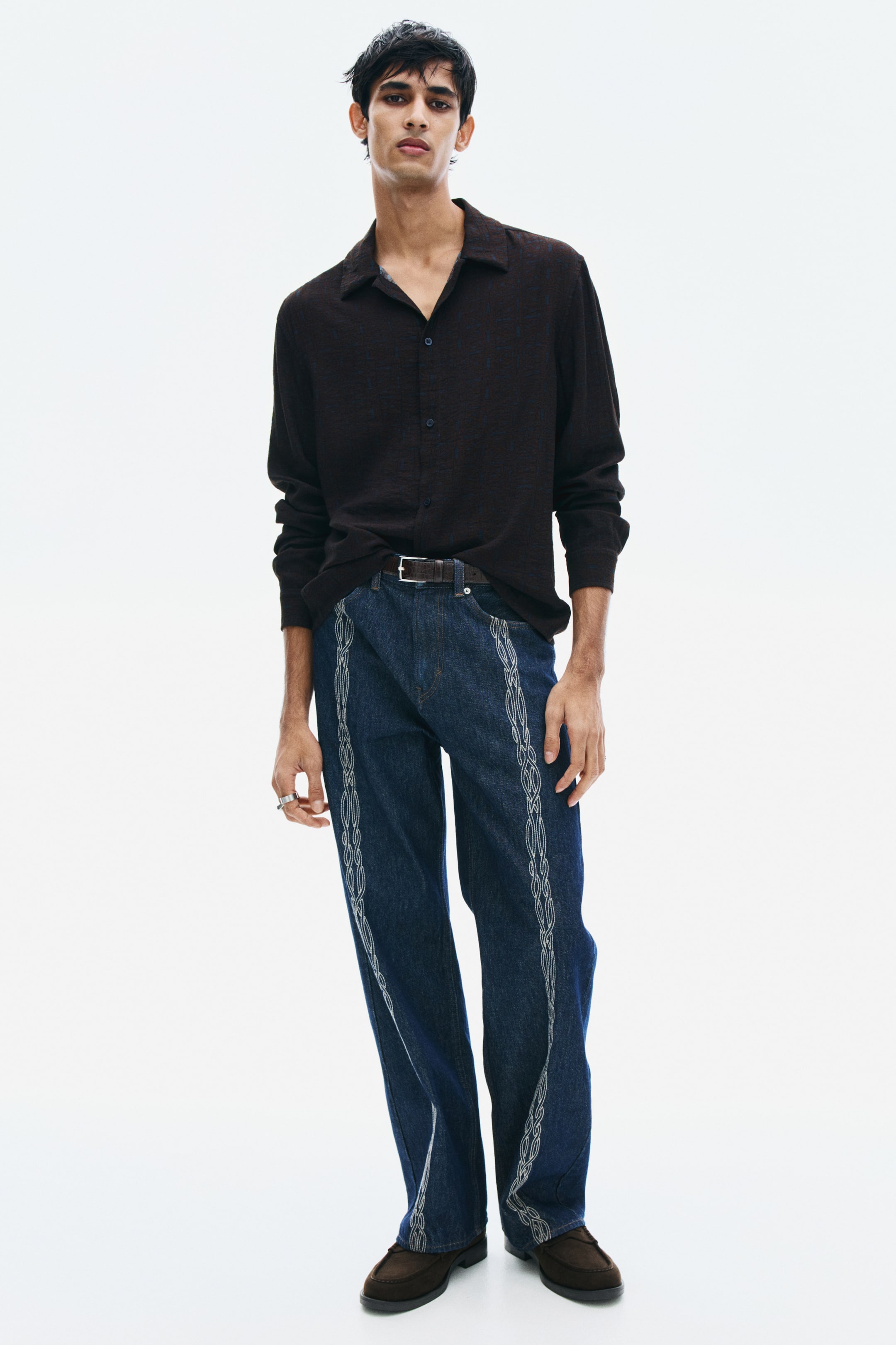 View larger image: The model is wearing a dark brown shirt in a textured viscose blend, featuring a relaxed fit, a collar, and a button-down front. The long-sleeved shirt is styled with a partial tuck into dark blue jeans, completed with a brown leather belt.
