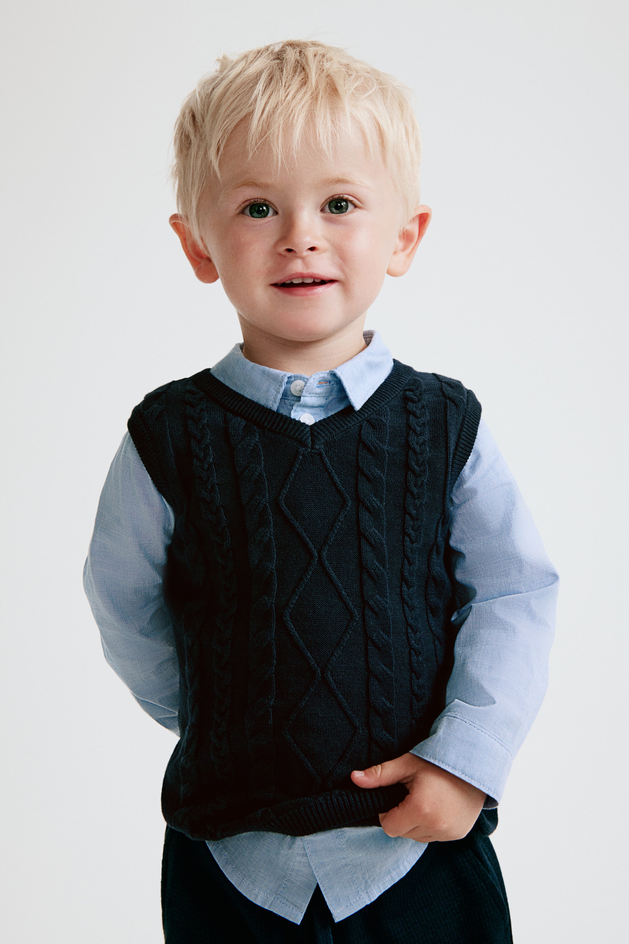 View larger image: A cotton set is shown from the front, featuring a light blue long-sleeved shirt layered underneath a dark navy blue cable-knit vest. The shirt is designed with a turn-down collar and a rounded hem extending below the vest, while the V-neck vest displays a diamond cable-knit pattern on the front and ribbed trims around the edges.