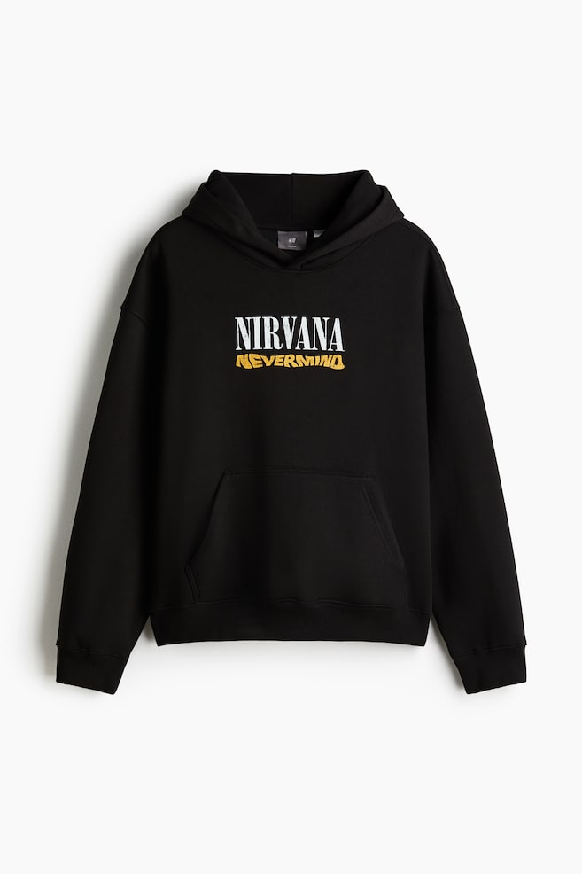 A black hoodie is shown from the front, featuring a loose fit with dropped shoulders and long sleeves. It has a lined hood, a kangaroo pocket, and ribbed cuffs and hem. The front is displaying "NIRVANA" in white text above "NEVERMIND" in distressed yellow text. - 1