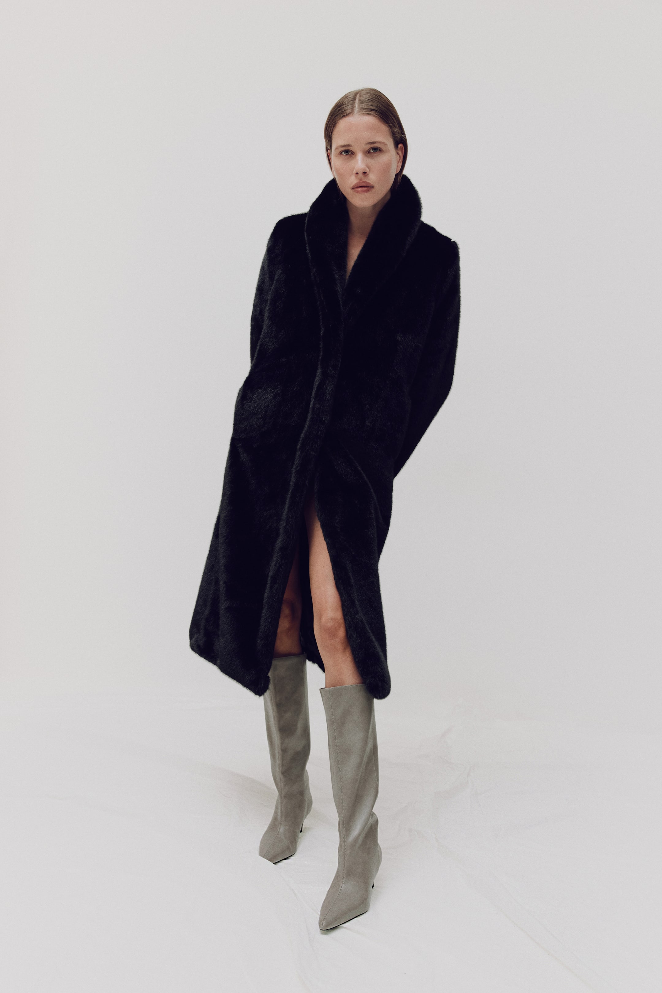 View larger image: A black faux fur coat is shown from the front, featuring a plush texture and a wide, stand-up collar. The long coat is worn open, creating a relaxed, flowing silhouette that falls below the knee.