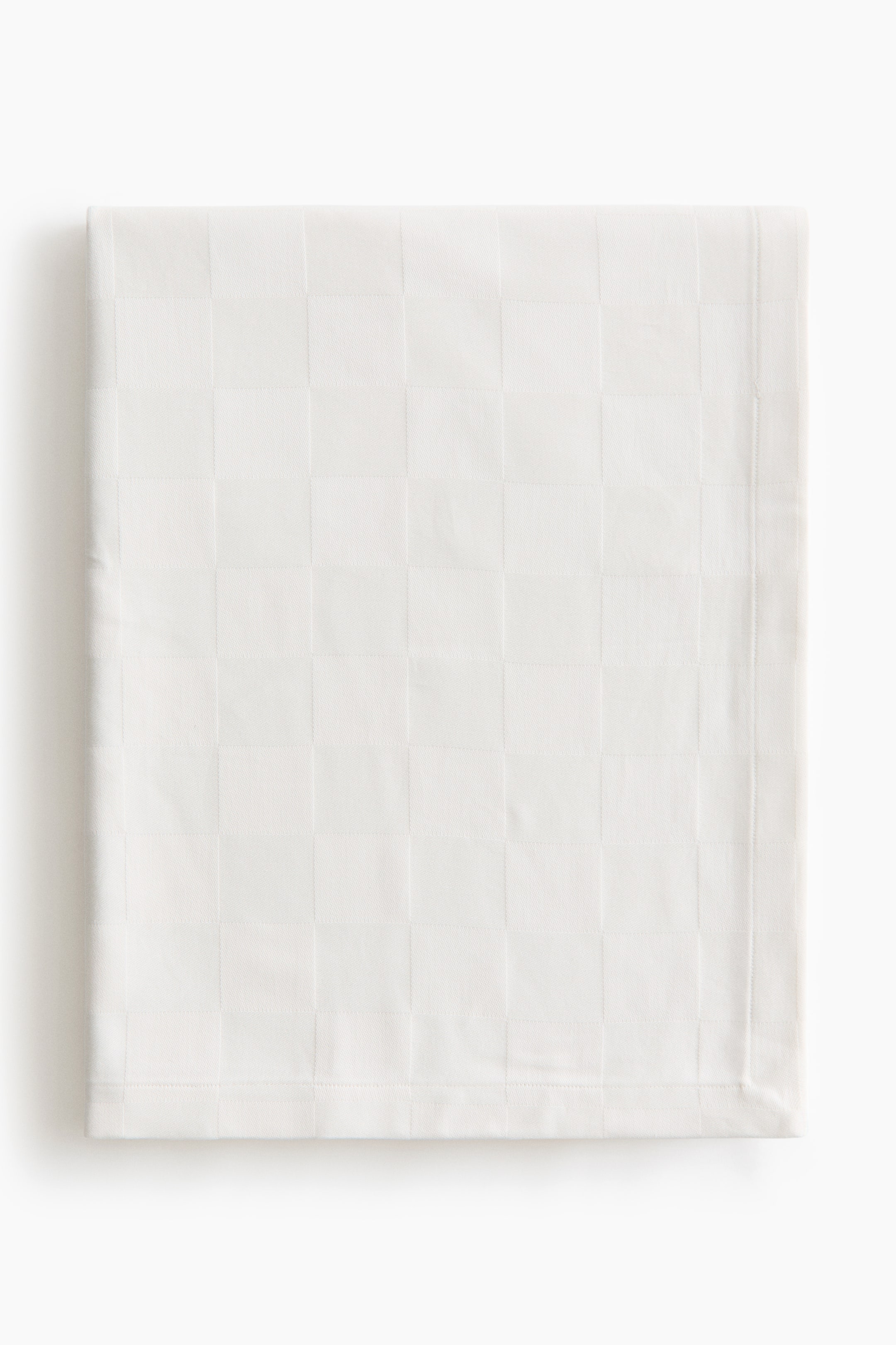 View larger image: A white cotton tablecloth is laid out folded flat and facing front, displaying a subtle, tone-on-tone woven checked pattern. The edges show a neat, wide folded hem with tonal stitching.