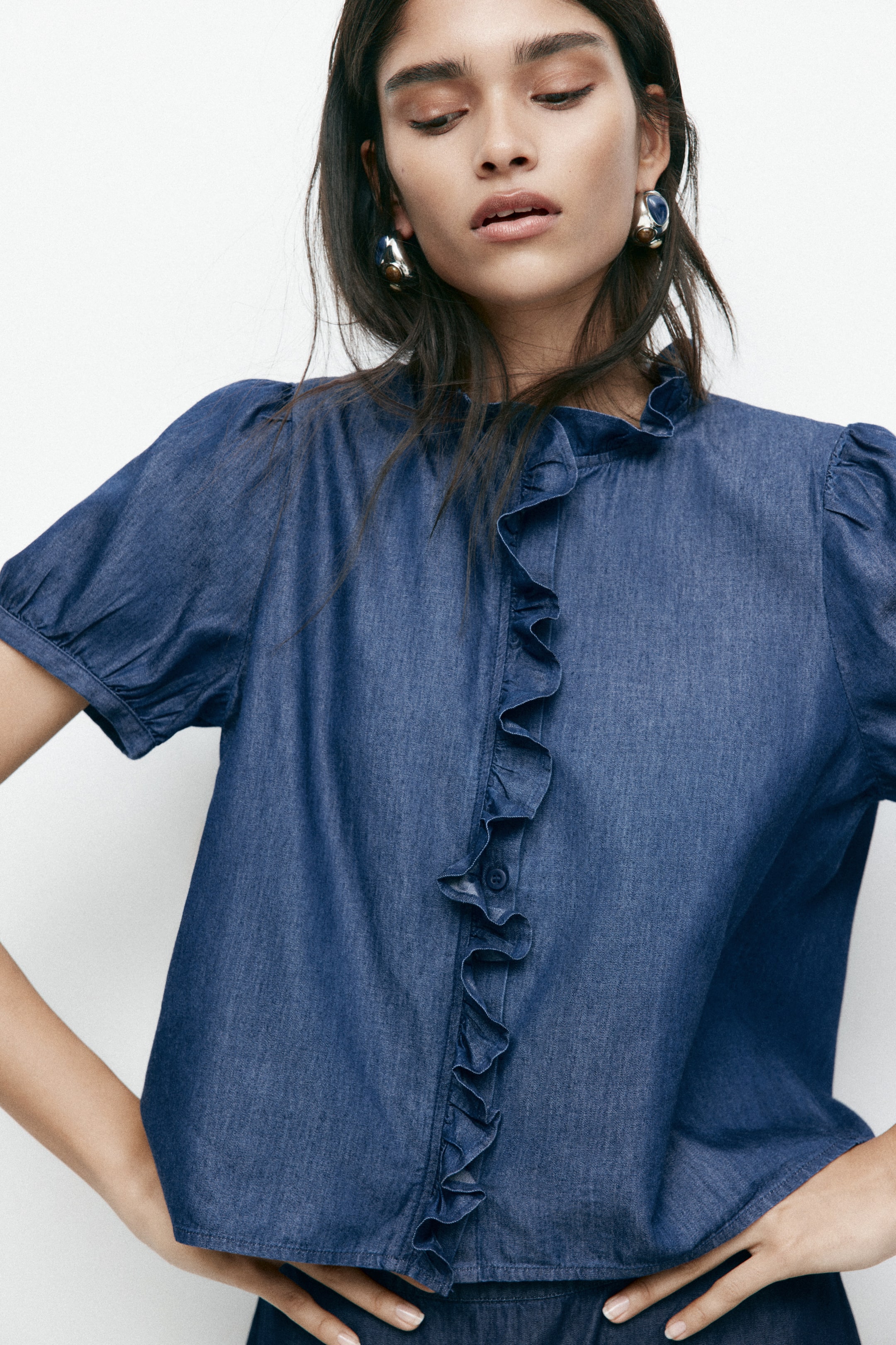 Visualizza immagine più grande: Una blusa in denim blu presenta un colletto arricciato a collo alto e una placca verticale con volant lungo la parte anteriore, che rivela una chiusura a bottoni. Sono visibili anche le maniche corte a sbuffo con polsini arricciati.