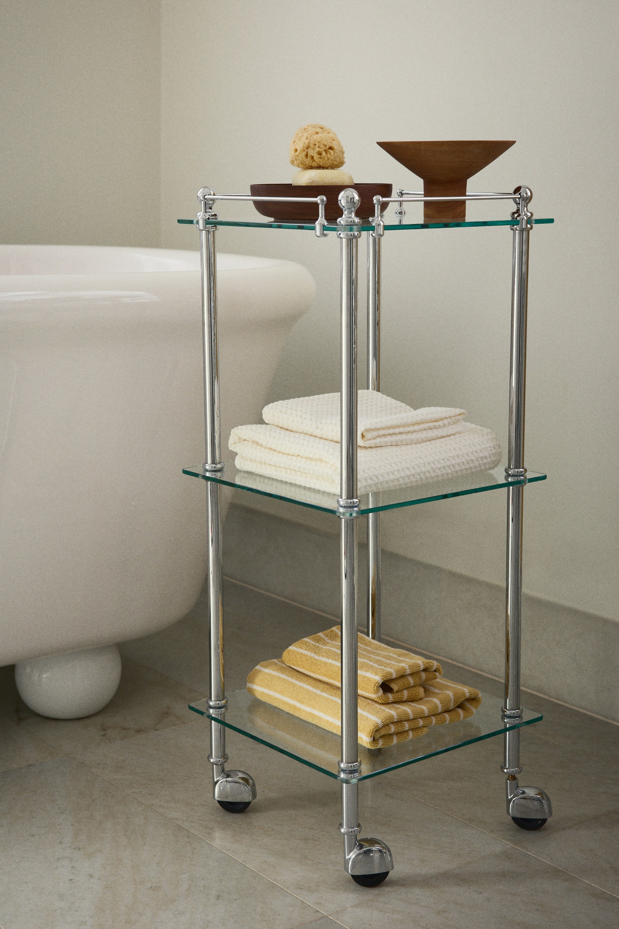 Visualizza immagine più grande: Un carrello a tre livelli cromato è posizionato su ruote, leggermente inclinato a sinistra. Presenta ripiani in vetro trasparente sostenuti da montanti cromati con terminali decorativi. I ripiani espongono asciugamani piegati e ciotole in legno con una spugna e sapone.