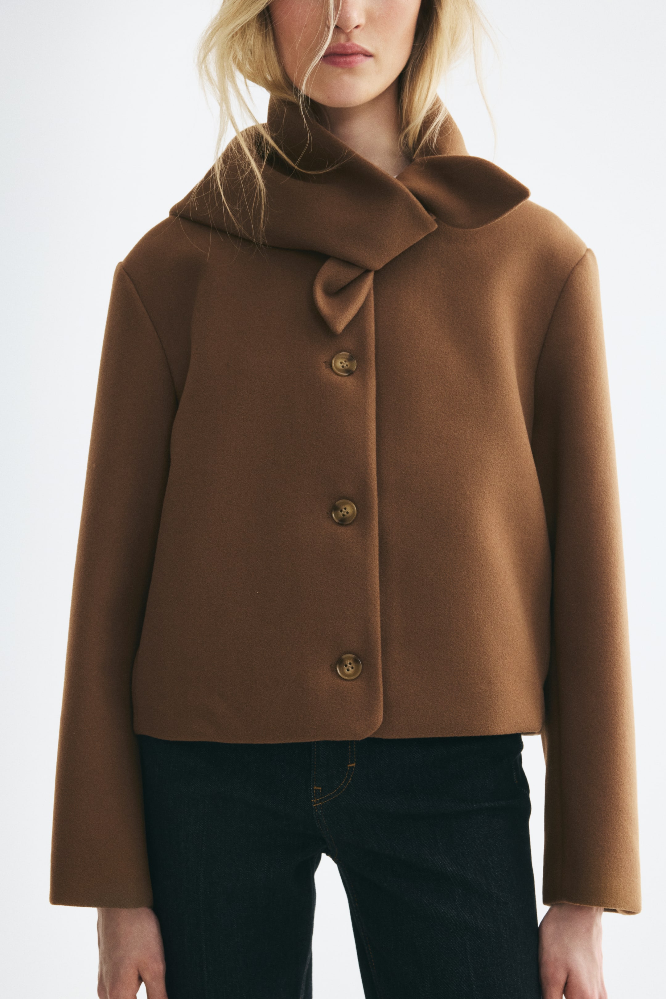View larger image: A short brown jacket in a soft, felted fabric is shown from the front, featuring an integrated draped scarf collar and a three-button closure down the front. The jacket has a boxy, cropped fit with long sleeves, ending just at the waistline of dark jeans.