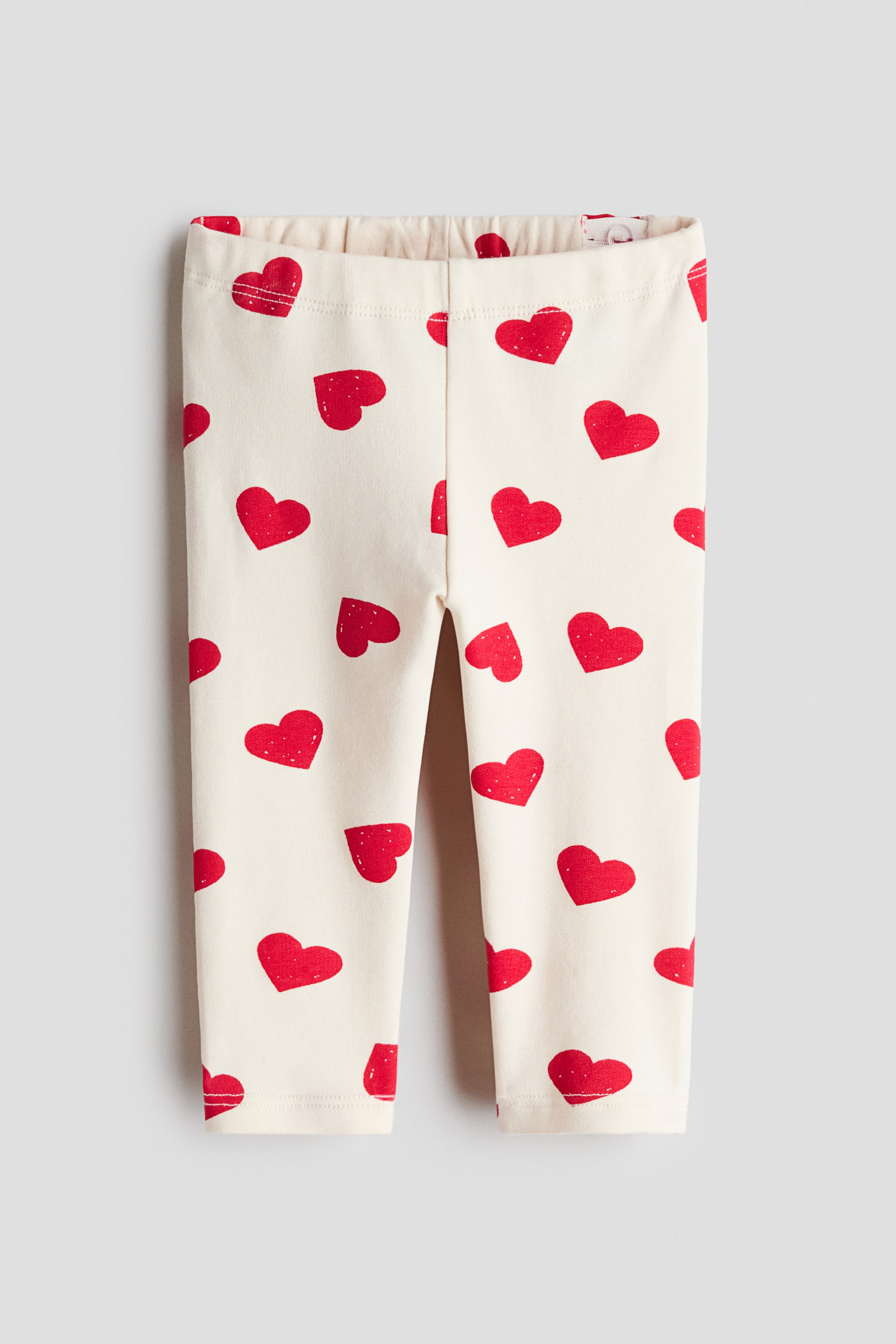 View larger image: Leggings are presenting flat, FRONT, in a cream-colored cotton jersey, patterned with scattered red hearts. An elasticated waistband is visible at the top, and the full-length legs are tapering towards the ankles.