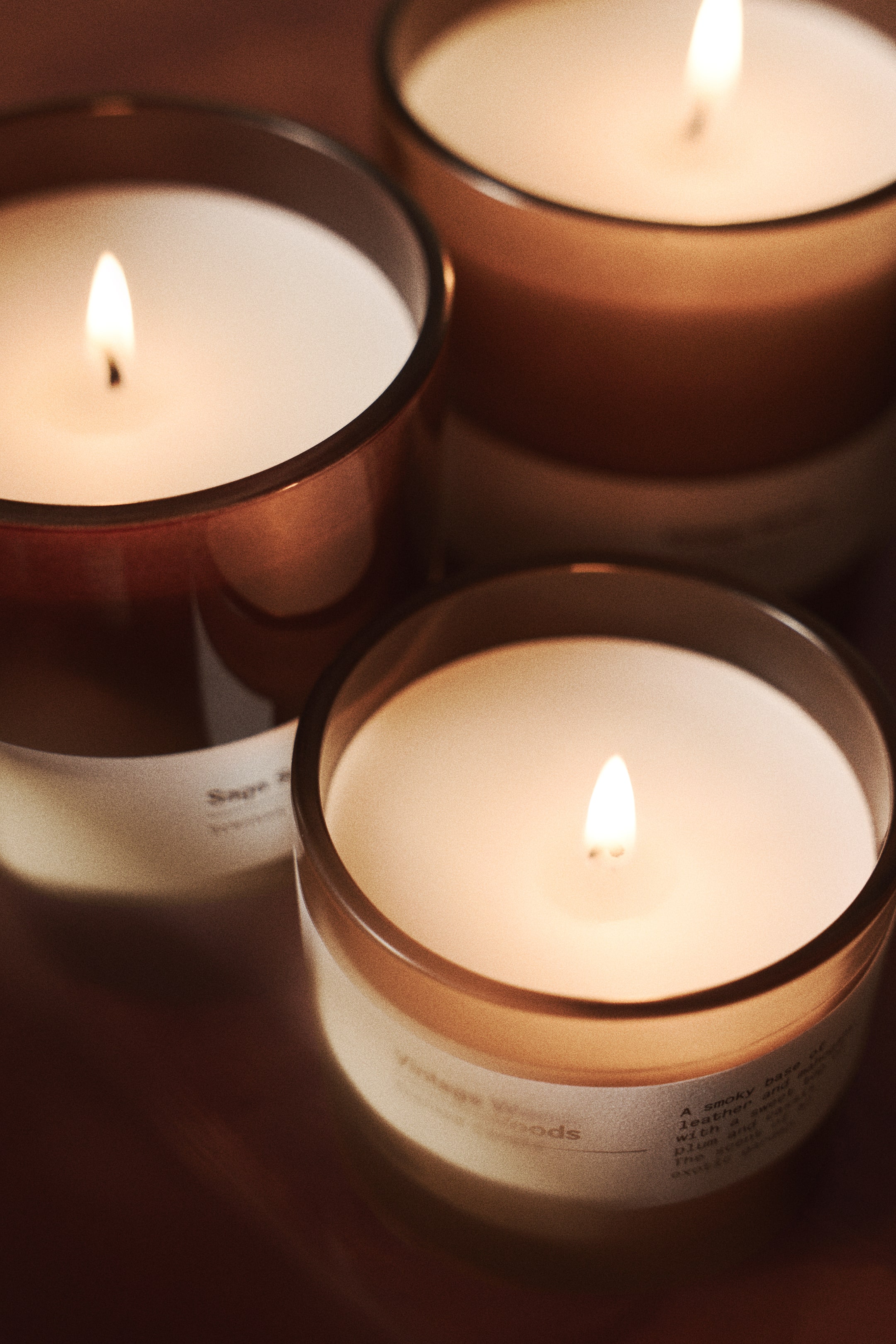 View larger image: Three candles in amber glass jars are burning, with each featuring a single lit wick casting a warm glow over the white wax. White paper labels are wrapped around the bottom of the jars, displaying printed text.