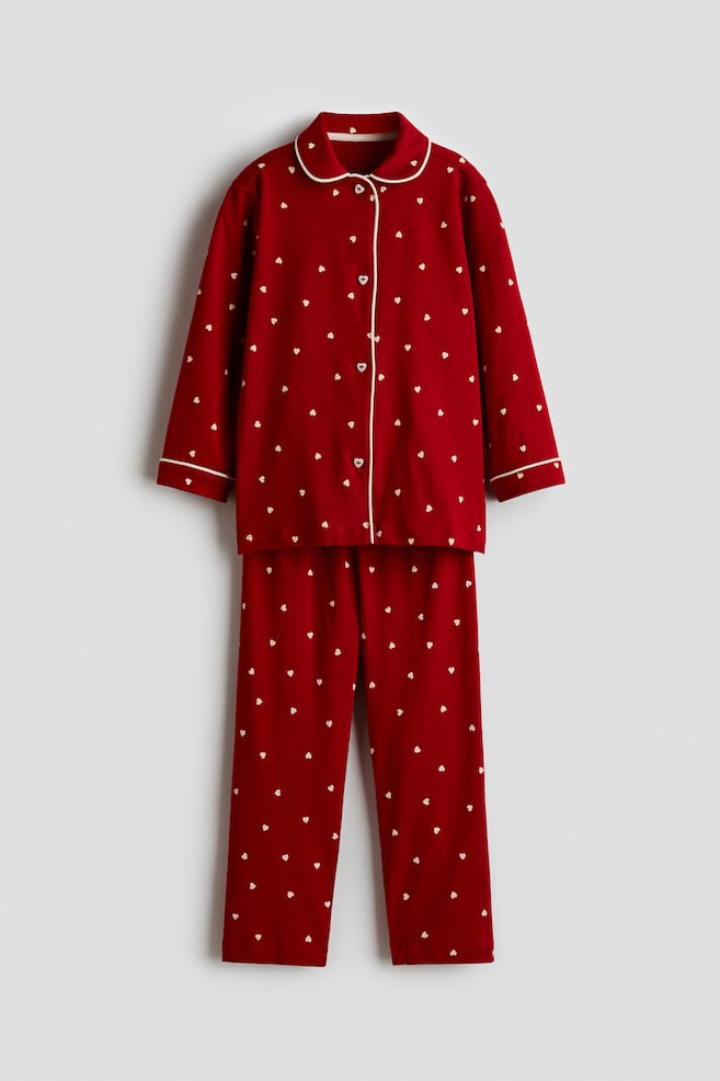 A red two-piece pajama set, front-facing, featuring an all-over pattern of small white hearts. The set includes a long-sleeved, button-up shirt with white piping along the rounded collar, placket, and cuffs, paired with matching straight-leg pants. - 1