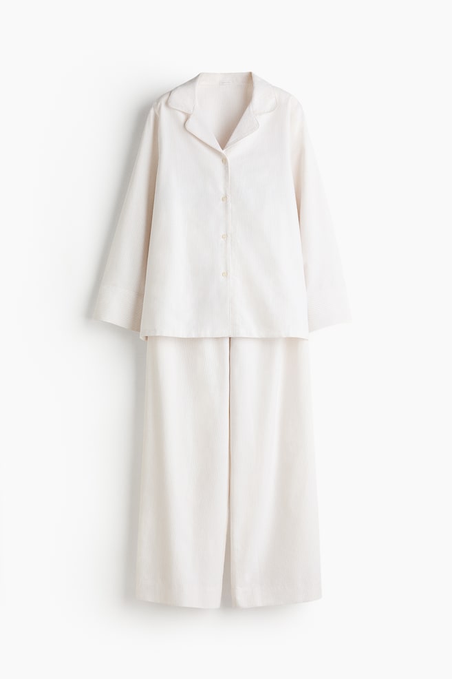 Un set pigiama bianco presenta una camicia a maniche lunghe con bottoni e pantaloni a gamba larga coordinati, entrambi caratterizzati da sottili righe verticali. La camicia è progettata con un colletto a revers e una chiusura frontale con quattro bottoni. - 1