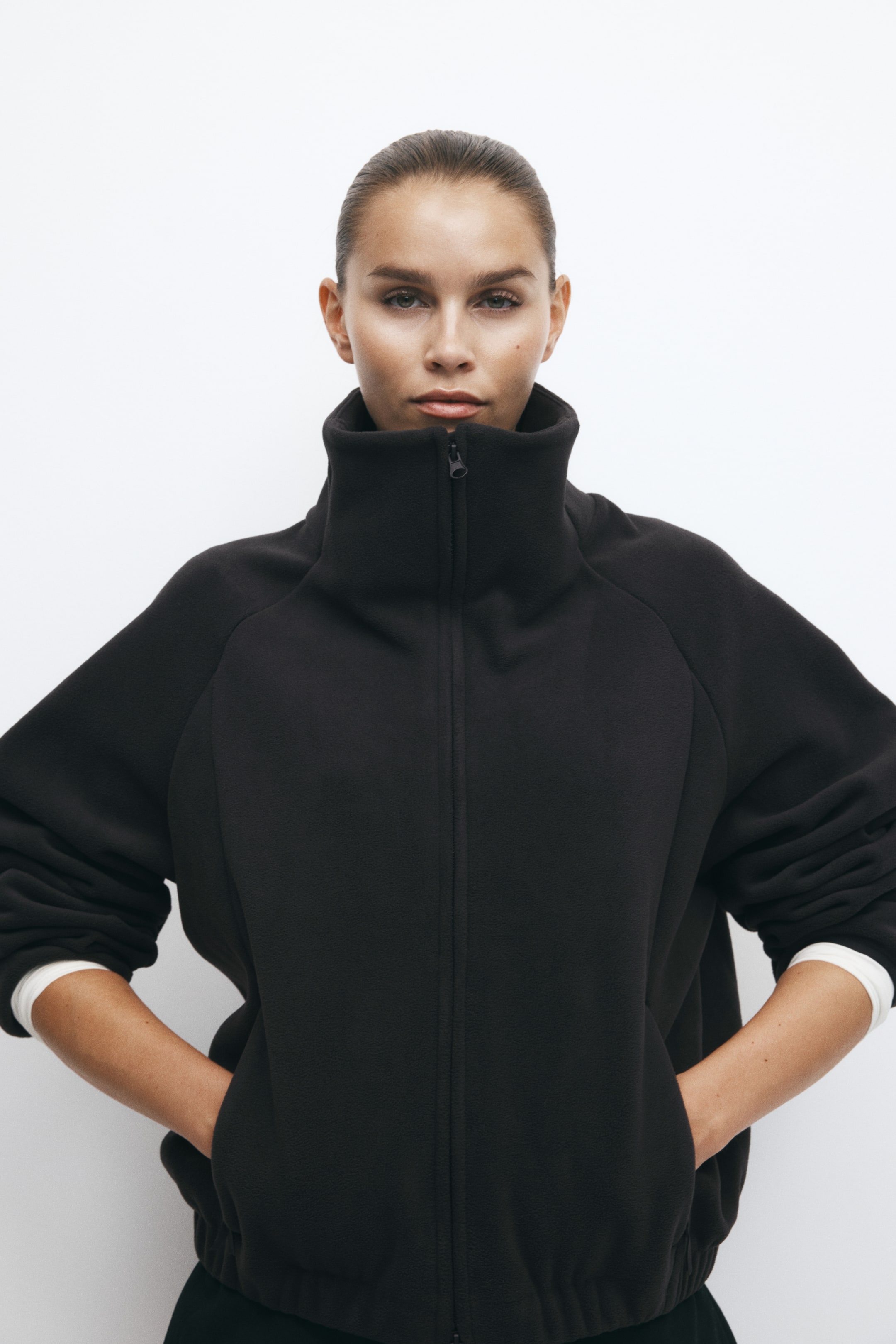 Vezi imaginea mai mare: O jachetă sport din fleece negru, cu guler înalt tip stand-up și închidere completă cu fermoar, este purtată de model. Modelul pozează cu mâinile introduse în buzunarele laterale, iar manșetele albe ale unui top cu mânecă lungă purtat în straturi sunt vizibile la încheieturi.