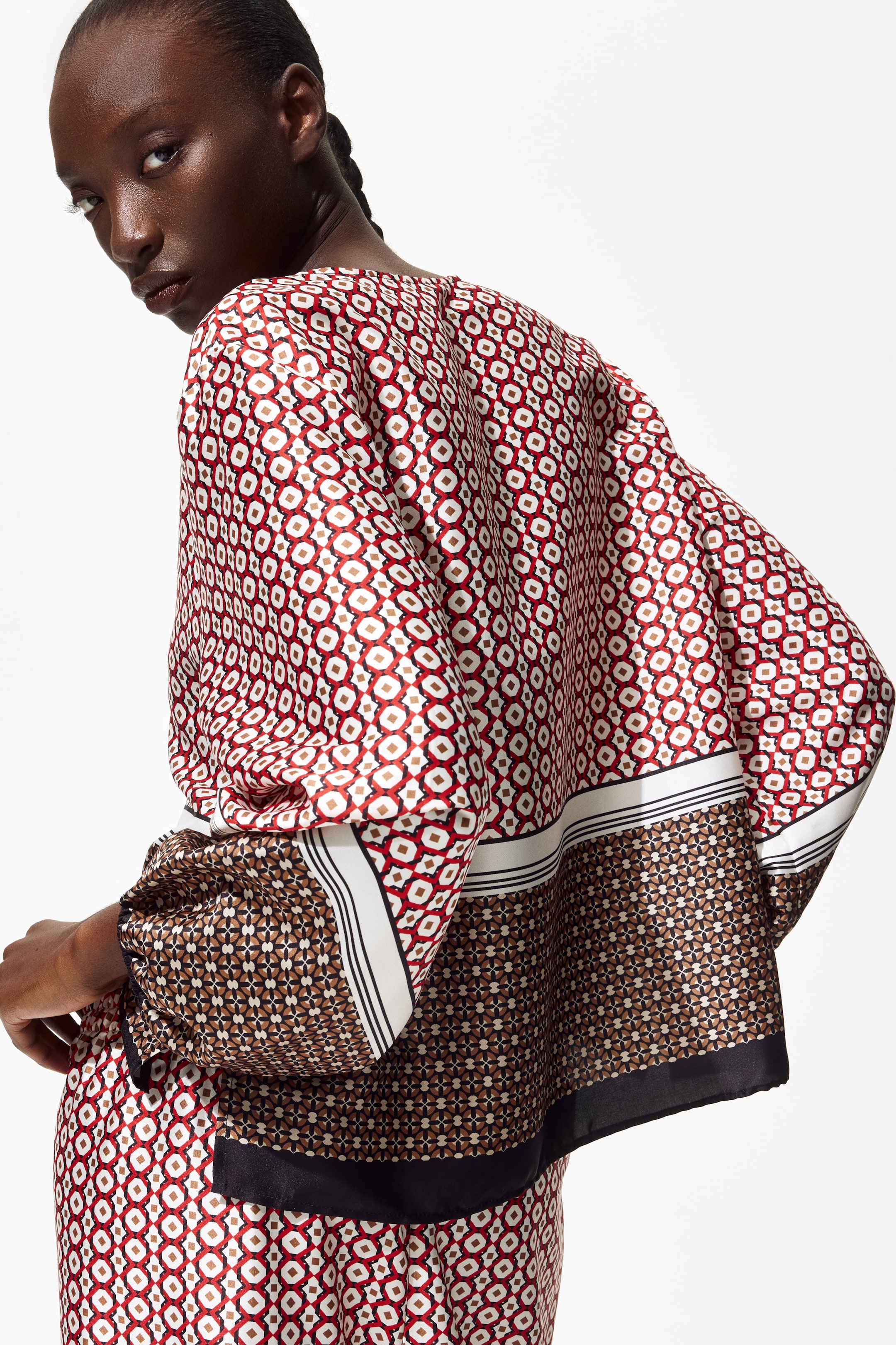 Visa större bild: En blus med avslappnad passform i vävt tyg med en lätt glans bärs av modellen. Den har långa ärmar och ett rött, vitt och brunt geometriskt mönster, med ett kontrasterande brunt och svart mönstrat band vid fållen, separerat av vita och svarta ränder. Modellen poserar med ena handen vilande nära midjan och bär även ett matchande mönstrat underplagg.