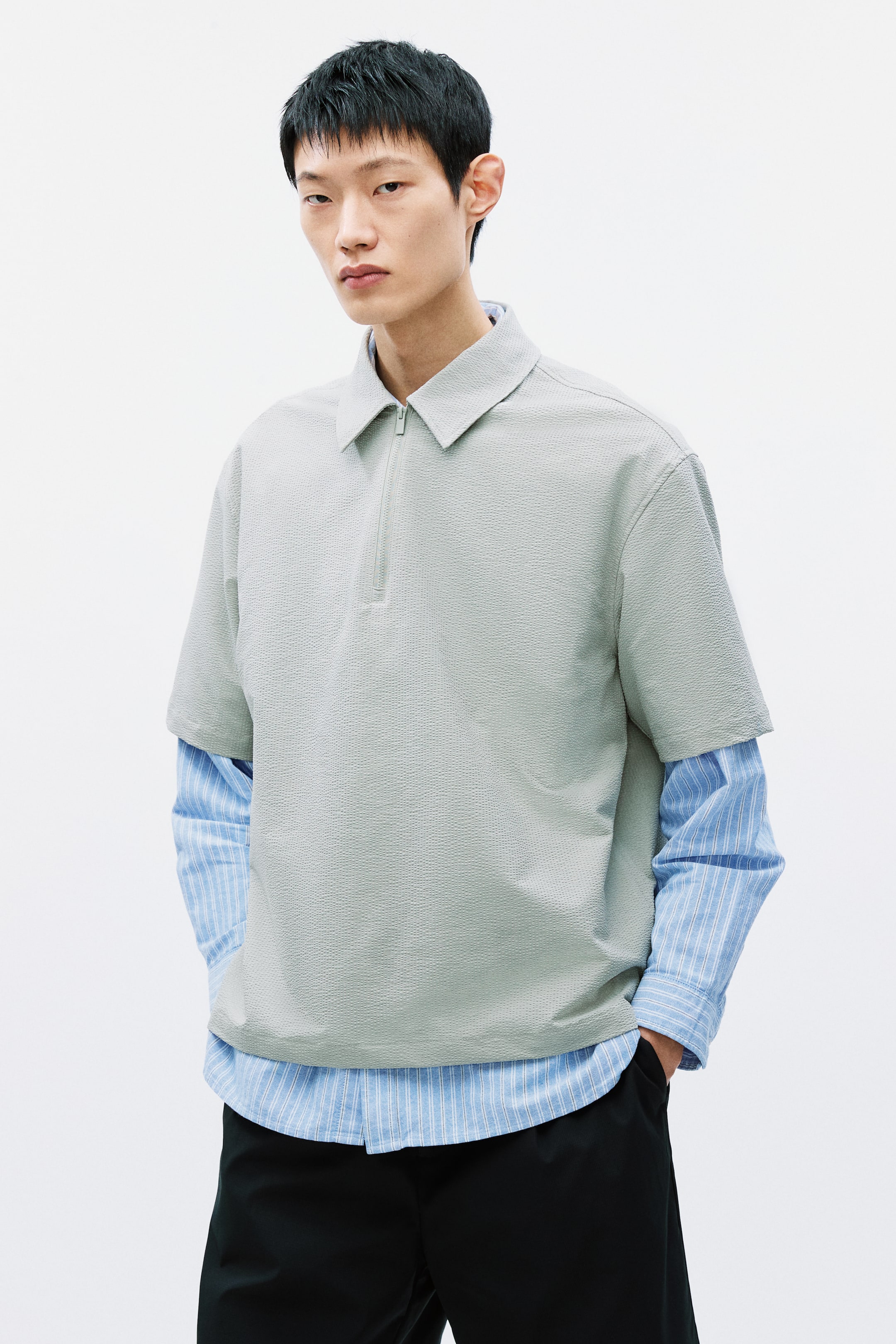 拡大画像を表示: ライトグリーンの半袖ポロシャツは、控えめなテクスチャーの生地、ポインテッドカラー、前面にクォータージップのプラケットが特徴です。ブルーとホワイトのストライプの長袖シャツの上に重ねて展示されています。