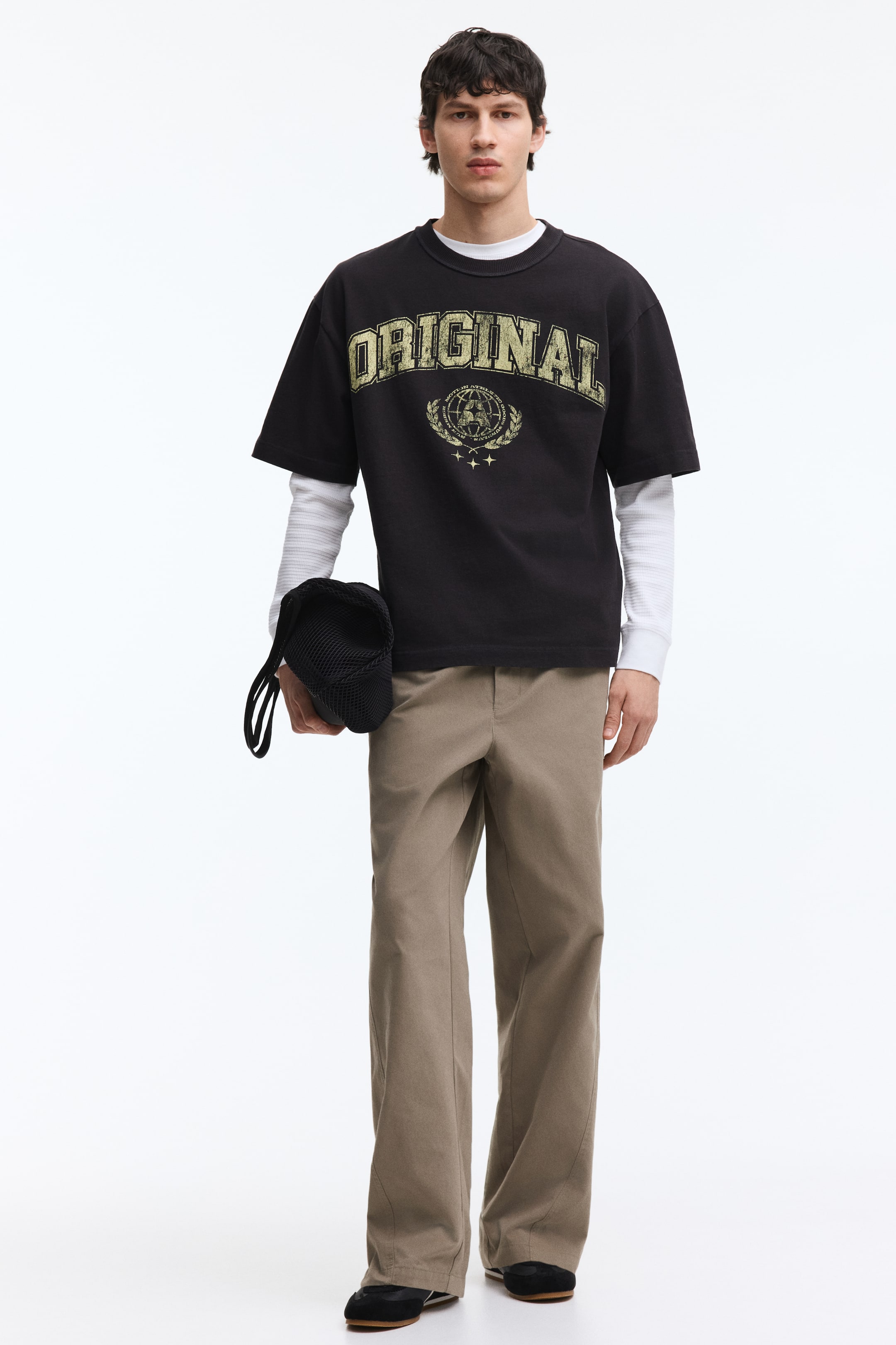 拡大画像を表示: 黒のオーバーサイズTシャツは、リブ付きのクルーネックと短くて幅広い袖が特徴です。前面には、淡い黄緑色の「ORIGINAL」という文字が、地球と月桂樹のグラフィックの上に重ねられて配置されています。白の長袖シャツの上にレイヤーされています。