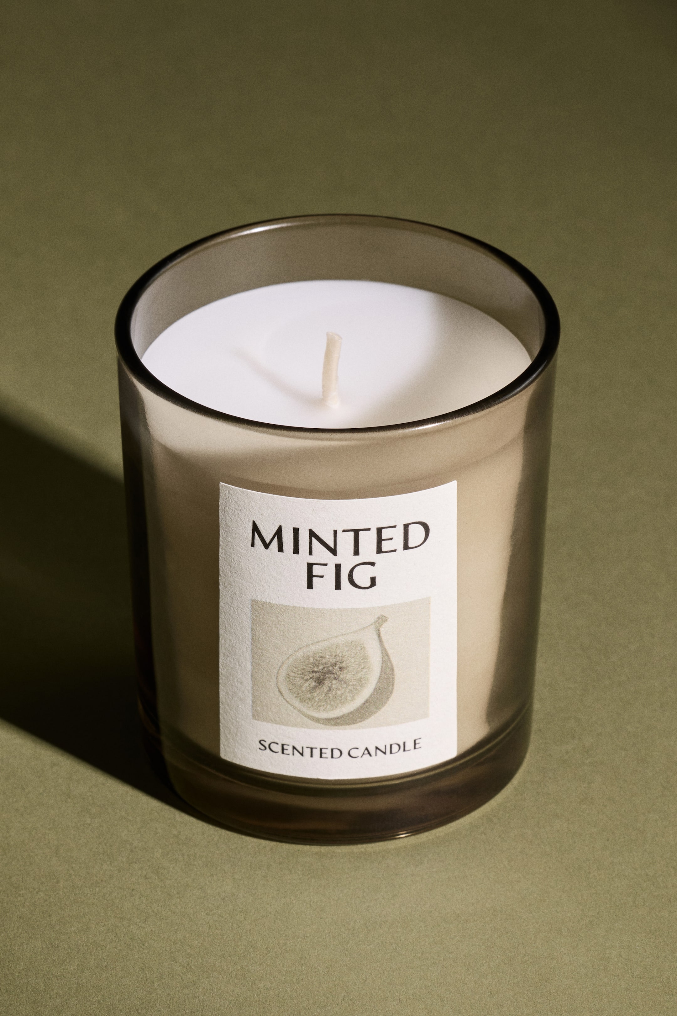 View larger image: A Minted Fig scented candle is visible, featuring a translucent dark grey glass jar filled with light beige wax and a white wick standing upright. A white rectangular label on the front displays the text "Minted Fig" and "Scented Candle", along with an illustration of a fig cross-section.